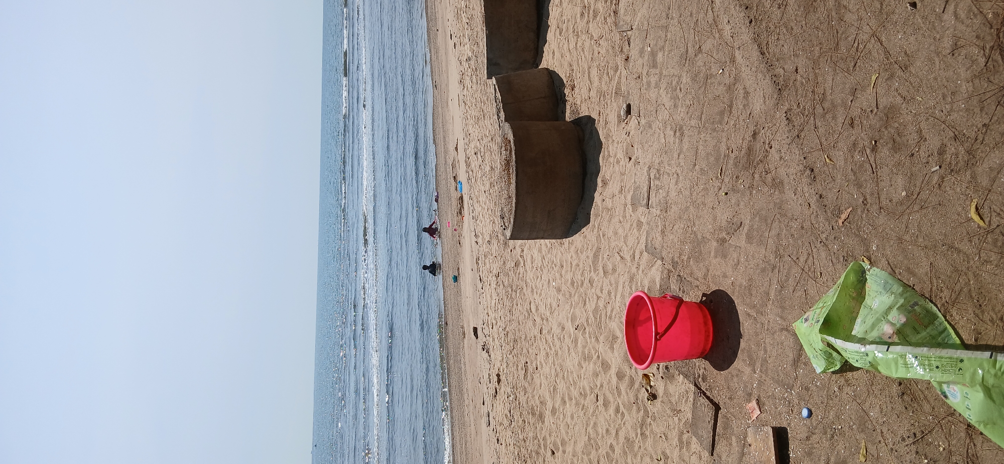 The image depicts a beach scene in Banyuglugur, Indonesia, under a clear sky. The foreground is dominated by sandy terrain with scattered debris, including a crumpled green plastic bag and a bright pink bucket. Several circular concrete structures are partially visible, suggesting man-made elements on the natural landscape. In the middle ground, two individuals, appearing to be children, are wading in the shallow water near the shoreline. The ocean stretches out in the background, with gentle waves reaching the beach and a distant haze obscuring the horizon. The overall atmosphere is one of a typical beach day, with recreational activities taking place.