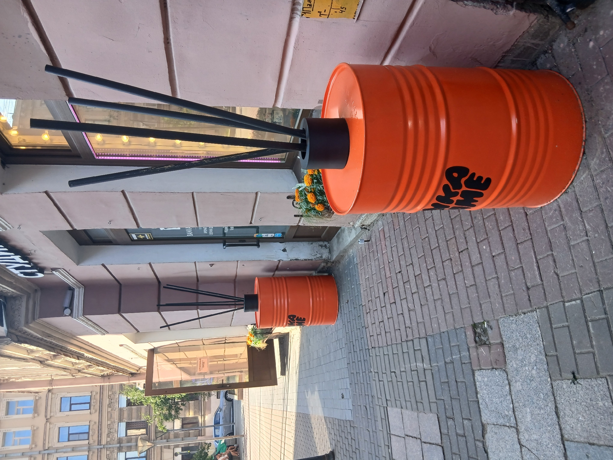 The image shows an outdoor scene on a paved sidewalk in front of a building. The primary subjects are two large, orange barrels, each with black rods extending from the top, resembling a large reed diffuser. One barrel is in the foreground, and another is further back. The barrel in the foreground has black text on it that appears to read "ME" and potentially other letters. Both barrels have some orange flowers placed around their base. The building behind the barrels has a light pink facade and large windows. Some of the windows have signs in a language that appears to be Cyrillic. One sign reads "СЛУЖЕБНЫЙ вход". The sidewalk is paved with interlocking bricks of various shades of gray and reddish-brown. In the background, there are views of other buildings, some with ornate architectural details and blue windows. The lighting suggests it is daytime, possibly late afternoon, with shadows cast across the pavement. No people are clearly visible in the foreground, though some figures might be present in the reflection of a window or further in the background. The overall impression is an urban streetscape with a quirky decorative element.