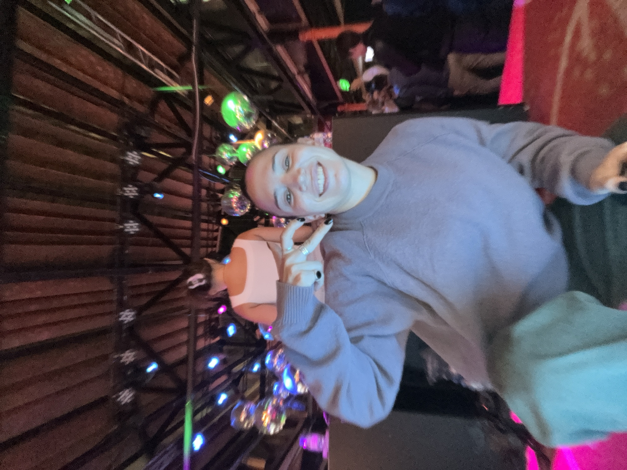 A tilted indoor shot captures a person, likely female, with a shaved head, wearing a light purple sweater and making a peace sign with their left hand. The person is smiling, their eyes looking into the camera. Behind them is a dimly lit interior space with disco balls hanging from what appears to be a metal frame structure, with multi-colored lights illuminating the area. The location and the setting indicate a party or entertainment venue, possibly a nightclub. No specific features allow for identifying the city as Moscow, Russia. Other people are present in the background, though they are out of focus and less discernible.