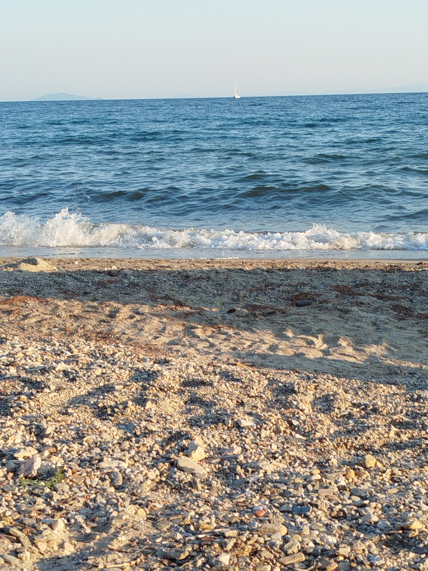 The image depicts a serene coastal scene in the Sithonia Municipal Unit, Greece. The foreground is a beach covered with small pebbles and sand, with shadows cast across the ground suggesting a bright day. The middle ground features a gentle wave breaking on the shore, creating a foamy white crest. Beyond the shoreline, the blue sea stretches to the horizon, calm with subtle ripples. In the distance, a white sailboat is visible on the water. Faint outlines of landmasses, likely islands or the mainland coast, can be seen on the far horizon under a clear, pale sky. There are no people or animals present in the frame.