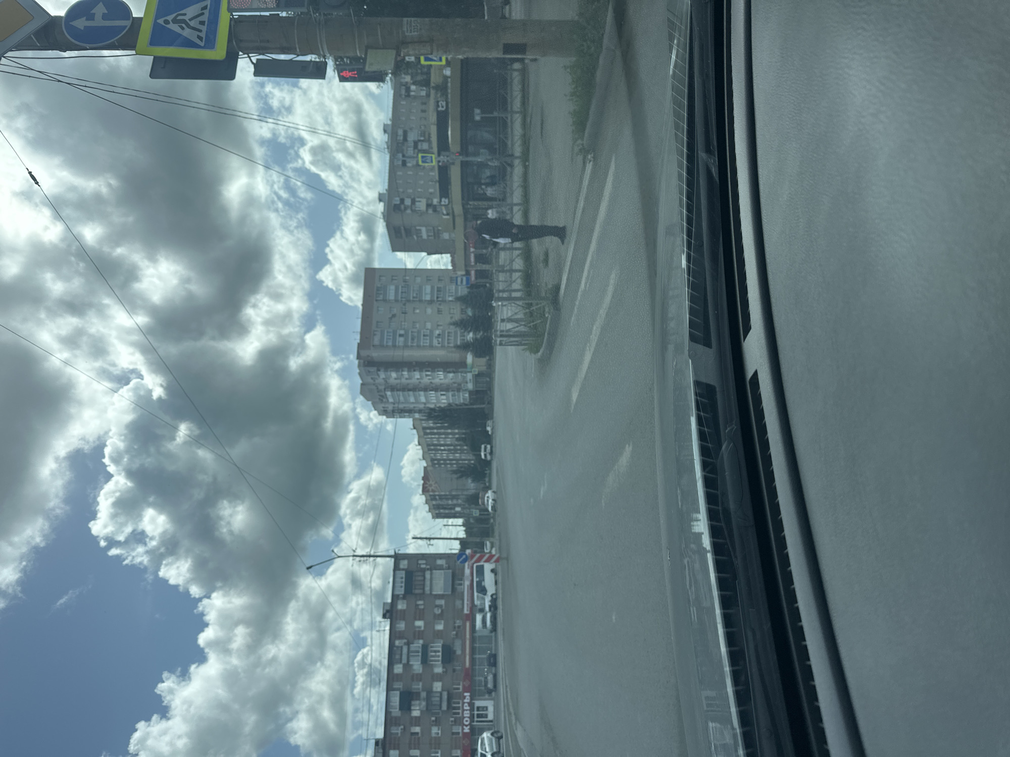 The image shows a view from a vehicle's dashboard, looking out onto a city street in Orsk, Russia, on a cloudy day. In the foreground, the vehicle's windshield wipers and part of the dashboard are visible. The street appears to be an intersection with multiple lanes of traffic. Several apartment buildings, typical of Soviet-era architecture, line the sides of the street. One building has a sign that reads "КОВРЫ", which translates to "Carpets". Traffic signs, including a pedestrian crossing sign and traffic lights, are present. A person is standing on the sidewalk near a crosswalk, looking at their phone. The sky is filled with dramatic, dark clouds, suggesting impending rain or a storm. Power lines are strung across the scene.