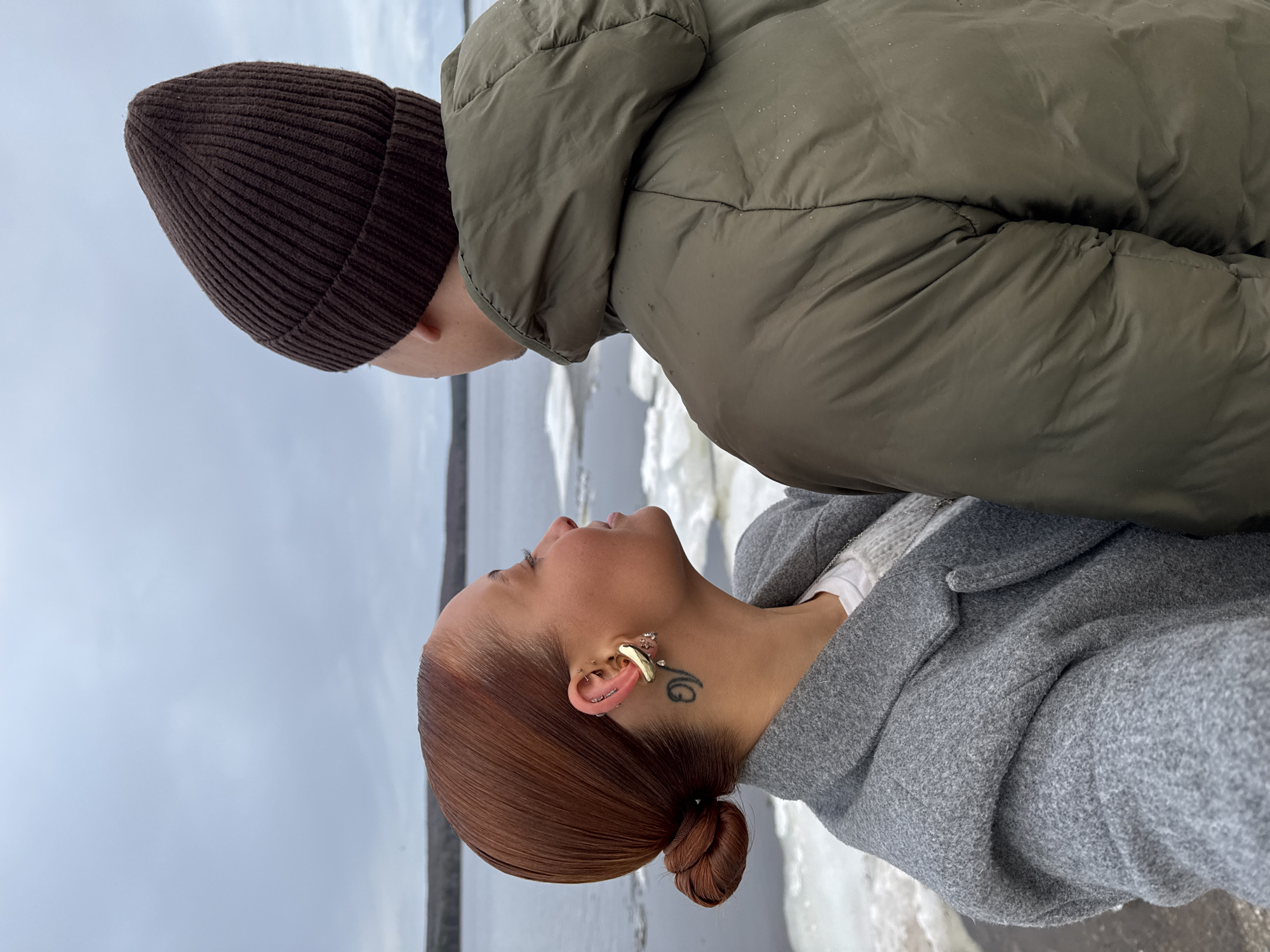 Two individuals, a man and a woman, are captured in an intimate, tender moment outdoors by a body of water, likely a river or lake, with visible chunks of ice. The image is framed as a close-up, focusing on their upper bodies.
The man, positioned on the left side of the frame, is largely obscured, with only his head and a portion of his back visible. He wears a dark brown knit beanie and an olive green puffer jacket, suggesting cold weather. He is leaning down towards the woman.
The woman, on the right, has reddish-brown hair pulled back into a neat, low bun. She is wearing a grey textured coat over what appears to be a white garment. Her head is tilted upwards, and she is looking towards the man, her expression conveying affection or anticipation. Notable details on the woman include multiple ear piercings, with a prominent gold-toned ear cuff or earring on her left ear, and a black tattoo of stylized script or a symbol on her neck just below her ear.
The background shows a tranquil, yet cold, environment. A grey, overcast sky meets the horizon of a large body of water, which is partially covered with floating ice. The diffused light suggests daytime, possibly a winter or early spring day. The cold weather is evident from the individuals' warm clothing and the icy water.
The overall scene depicts a private, affectionate interaction between the two people amidst a serene, chilly landscape. Given the presence of ice and warm attire, the location of Chaikovsky, Russia, aligns with the environmental cues in the image.