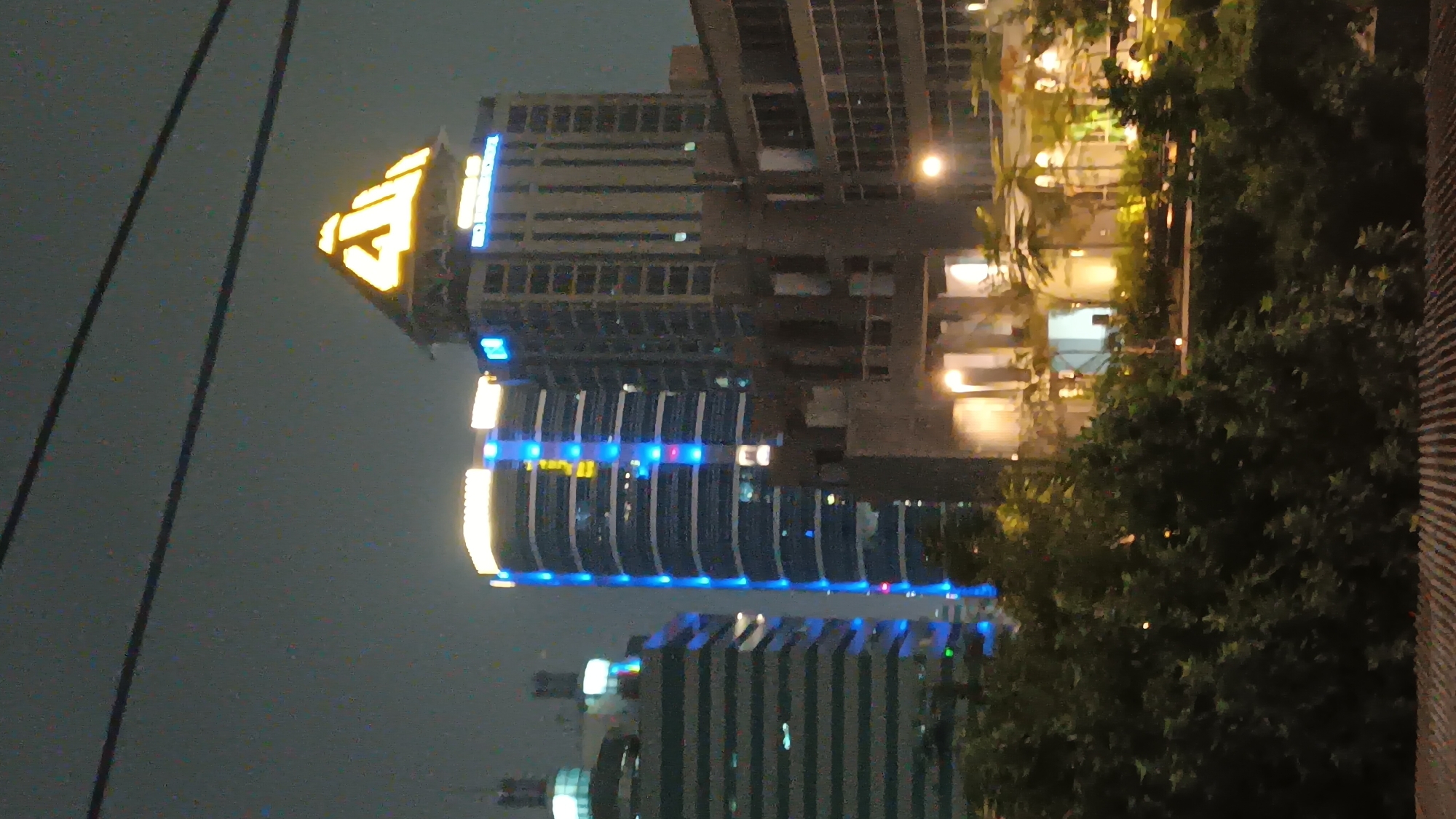 This image shows a nighttime view of several modern buildings in South Jakarta, Indonesia. The most prominent structure is a tall building with a pyramid-shaped top illuminated with golden light, displaying Arabic calligraphy. To its left, another tall, cylindrical building is adorned with vertical blue lights. In the foreground, lush green trees partially obscure the view of the lower levels of the buildings, suggesting a natural element integrated into the urban landscape. The sky is dark, indicating it is nighttime. There are no people or discernible actions visible in the image. The overall scene depicts an illuminated cityscape with a focus on architectural lighting.
