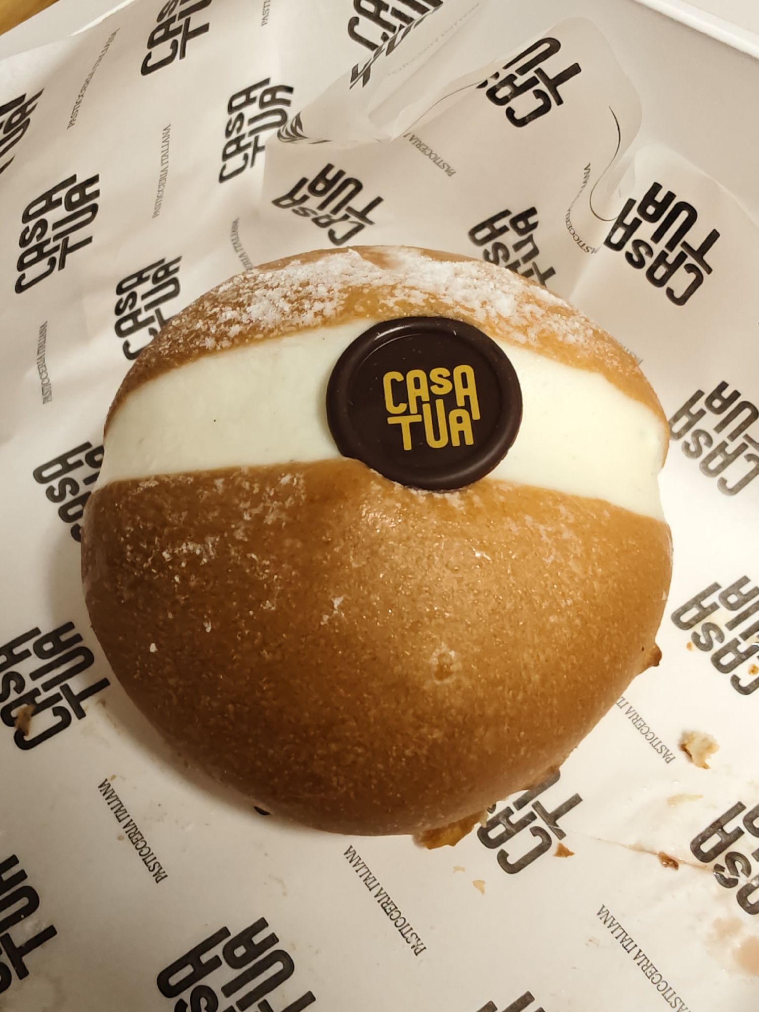 The image shows a close-up, top-down view of a dessert, likely a brioche bun filled with cream, resting on white paper with a repeated pattern of "CASA TUA" and "PASTICCERIA ITALIANA". The dessert is golden brown and dusted with powdered sugar. A circular chocolate disc with the "CASA TUA" logo in yellow is embedded in the cream filling. The paper wrapping is slightly crumpled. The scene appears to be indoors, possibly a bakery or cafe, and no specific location within Petah Tikva, Israel, can be determined from the image alone.