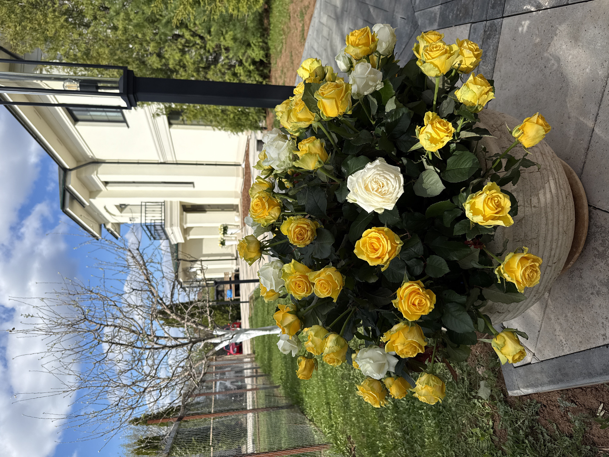 The image shows a bouquet of yellow and white roses in a stone pot outdoors. The location is likely a residential area in the city of Гольяны, Russia, as evidenced by the architecture of the houses in the background. A black lamppost stands beside the bouquet. In the background is a house, the sky and a bare tree behind a wire fence. The flowers are in full bloom, suggesting a warm season.