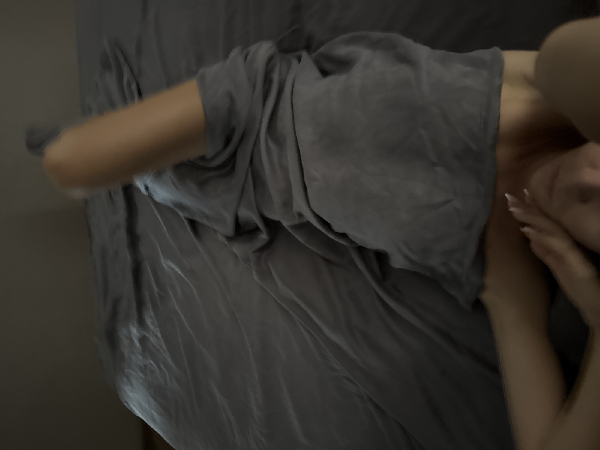A top-down, blurry shot shows a person lying on a bed with gray sheets. The person is partially covered by the sheets, with their arm and shoulder visible. Their skin tone is light, and they have manicured fingernails. The person's head is turned, and their face is mostly obscured by their hair and hand, but it appears they are looking downwards with their eyes closed and a slight smile. The lighting is dim, suggesting it could be nighttime or a dimly lit room. There is no visible text in the image.