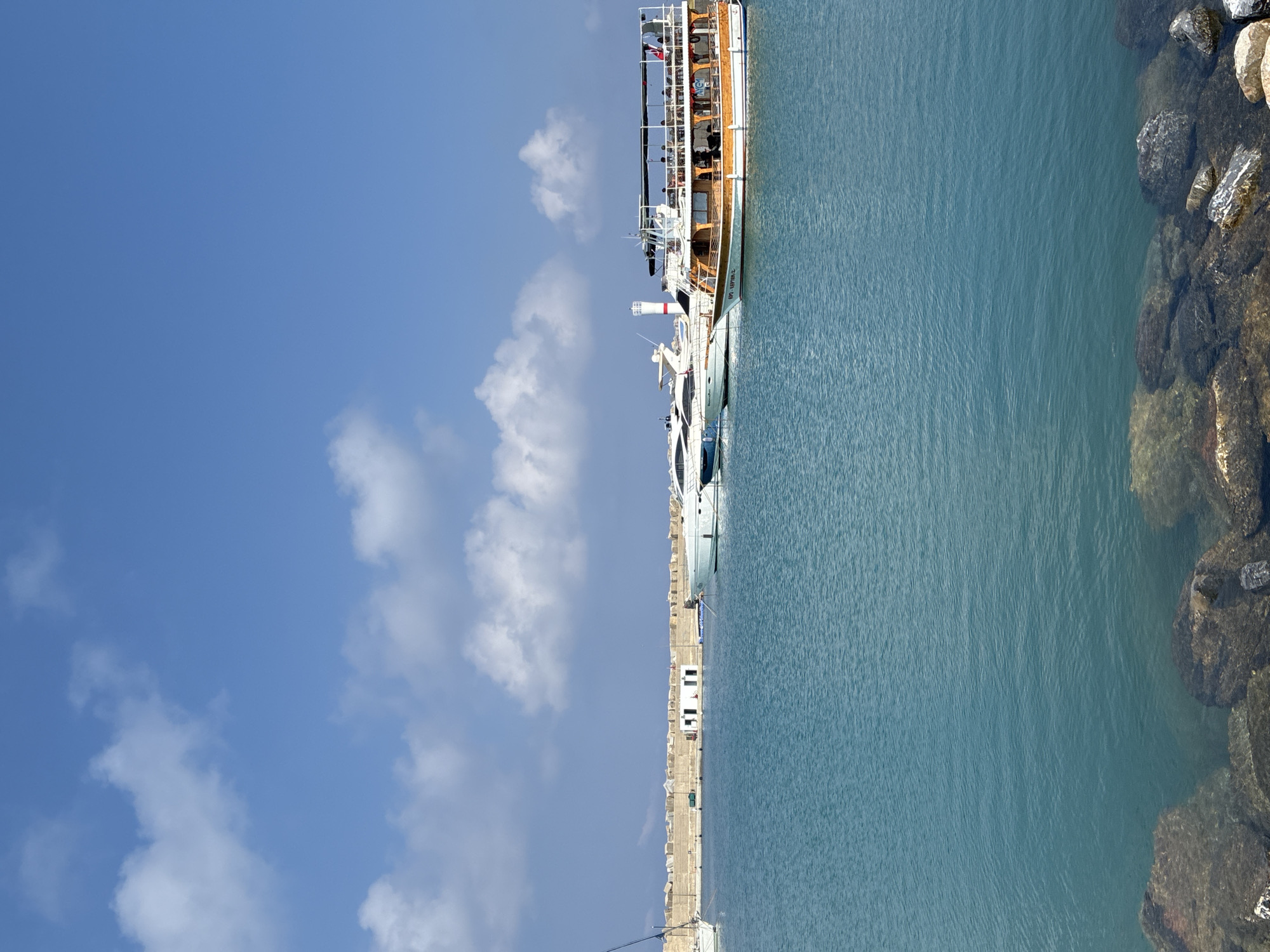 This is a photograph taken in the city of Gazipaşa, Turkey, likely at its harbor. The scene depicts a clear blue sky with scattered white clouds. In the foreground, the turquoise waters of the sea are visible, with rocks lining the bottom edge of the frame. Several boats are moored in the harbor. On the left, a sailboat's mast is partially visible. In the middle ground, two white luxury yachts are docked. To the right, a larger, multi-decked tour boat with a wooden lower section and a covered upper deck is prominent. The boat appears to be carrying passengers, some of whom are visible on the upper deck. In the background, a long, stone breakwater with several small white structures on top runs horizontally across the image, separating the harbor from the open sea. A white lighthouse with a red stripe is situated behind the yachts. The overall impression is a sunny day at a harbor with boats and calm waters.