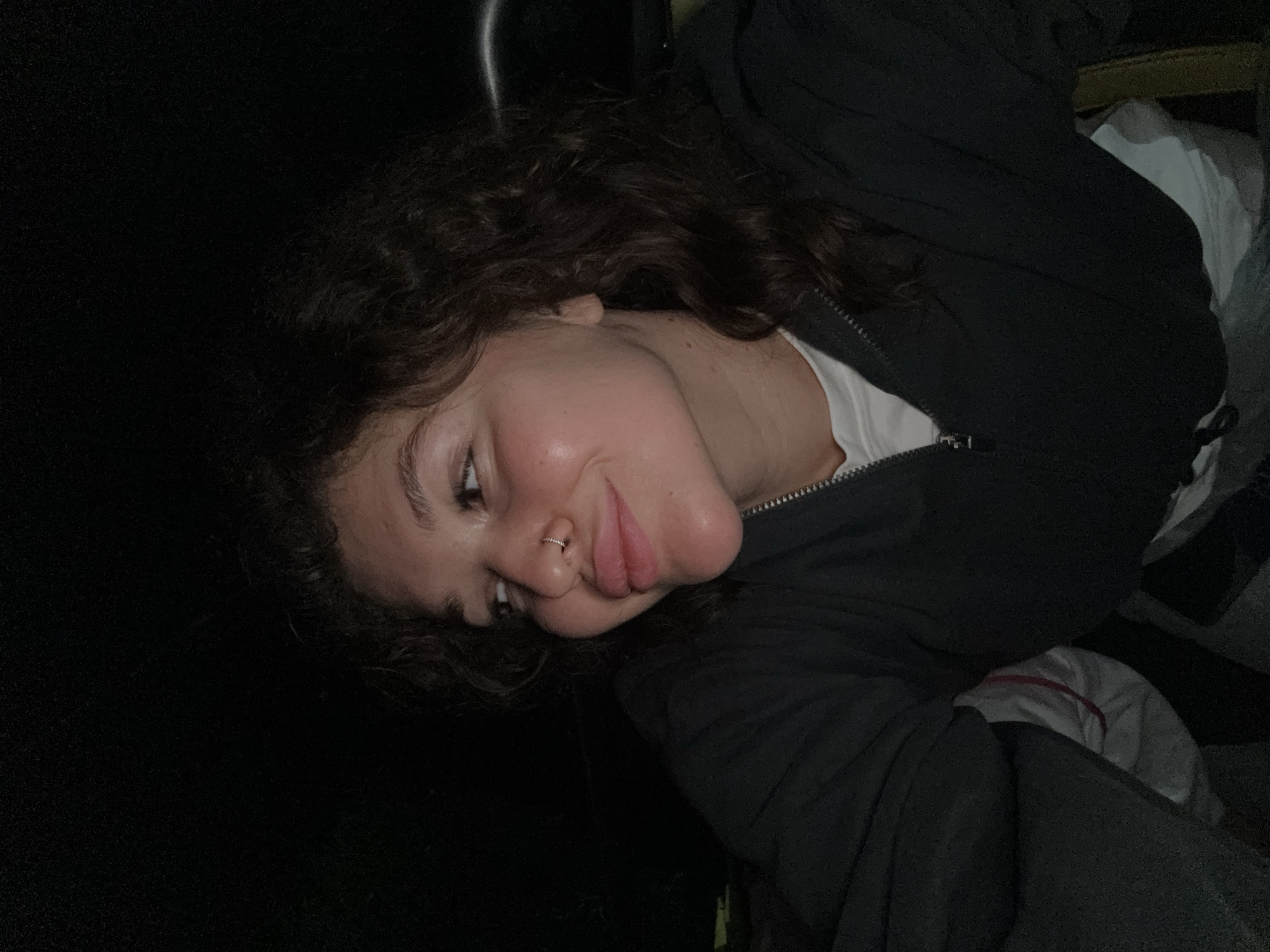 This close-up portrait features a young woman captured in what appears to be low-light or nighttime conditions. The image is oriented with the woman's head on the left side, her body extending to the right, and is rotated 90 degrees clockwise. She has dark, curly hair and fair skin, with a slight flush on her cheeks. A small, silver hoop nose ring is visible on her left nostril.
The woman is looking towards the left side of the frame with a subtle smile or smirk, her lips slightly puckered. She is wearing a dark, possibly black, zip-up jacket over a white top, with the jacket's zipper visible down the front. The background is largely dark, almost black, with some indistinct lighter areas, including white and grey fabric or objects, visible on the right side of the frame, suggesting an indoor or sheltered setting. The lighting highlights her face against the dark surroundings.
No specific activity is taking place beyond posing for the photo. The time of day is inferred as nighttime due to the darkness. There are no weather cues or visible text. Although not visually discernible in the image, the provided context indicates the location is Kintamani, Indonesia. The emotion conveyed is one of casualness or mild amusement.