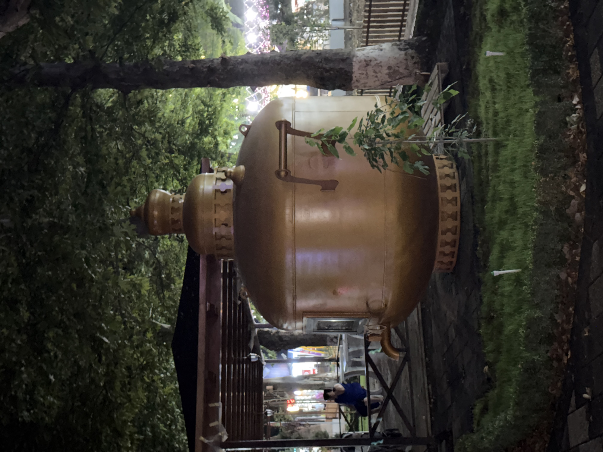 A large, golden samovar is the central object in this image, positioned outdoors under a tree at dusk or night. The samovar is ornate, with decorative carvings and handles. A small plant with green leaves grows near the top of the samovar. Behind the samovar, a wooden fence and a grassy area are visible, with some fallen leaves on the ground. In the background, there are lights, suggesting the presence of buildings or a street. A person wearing a blue shirt and dark pants is seen bending down in the lower right portion of the image, near a metal structure that appears to be part of a seating area or gazebo. The overall impression is a nighttime scene in a public outdoor space, likely a park or garden, with a prominent cultural artifact. The image is oriented vertically, rotated 90 degrees clockwise.
