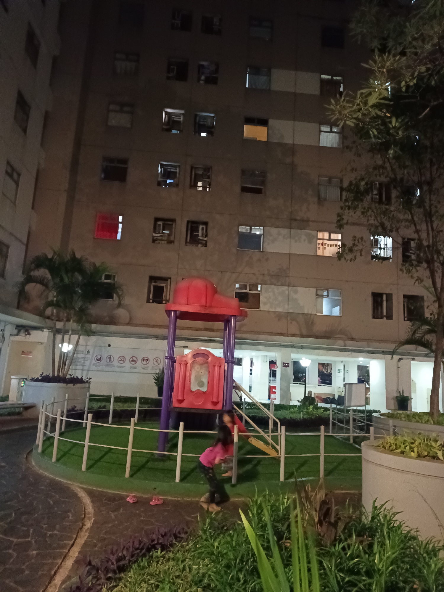 A nighttime outdoor scene in a residential complex in South Jakarta, Indonesia, shows a multi-story building with numerous windows, some illuminated from within. In the foreground, a playground structure with red and purple elements is visible. A young person, wearing a pink shirt and dark pants, is interacting with the playground, appearing to be sliding down a yellow slide. Pink flip-flops are placed on the paved area near the playground. The ground features a dark, patterned pathway and green artificial turf surrounding the play area. Lush green plants, including palm trees, are situated in large planters and along the edges of the scene. The overall environment suggests a common area within a condominium or apartment complex, illuminated by artificial lighting.