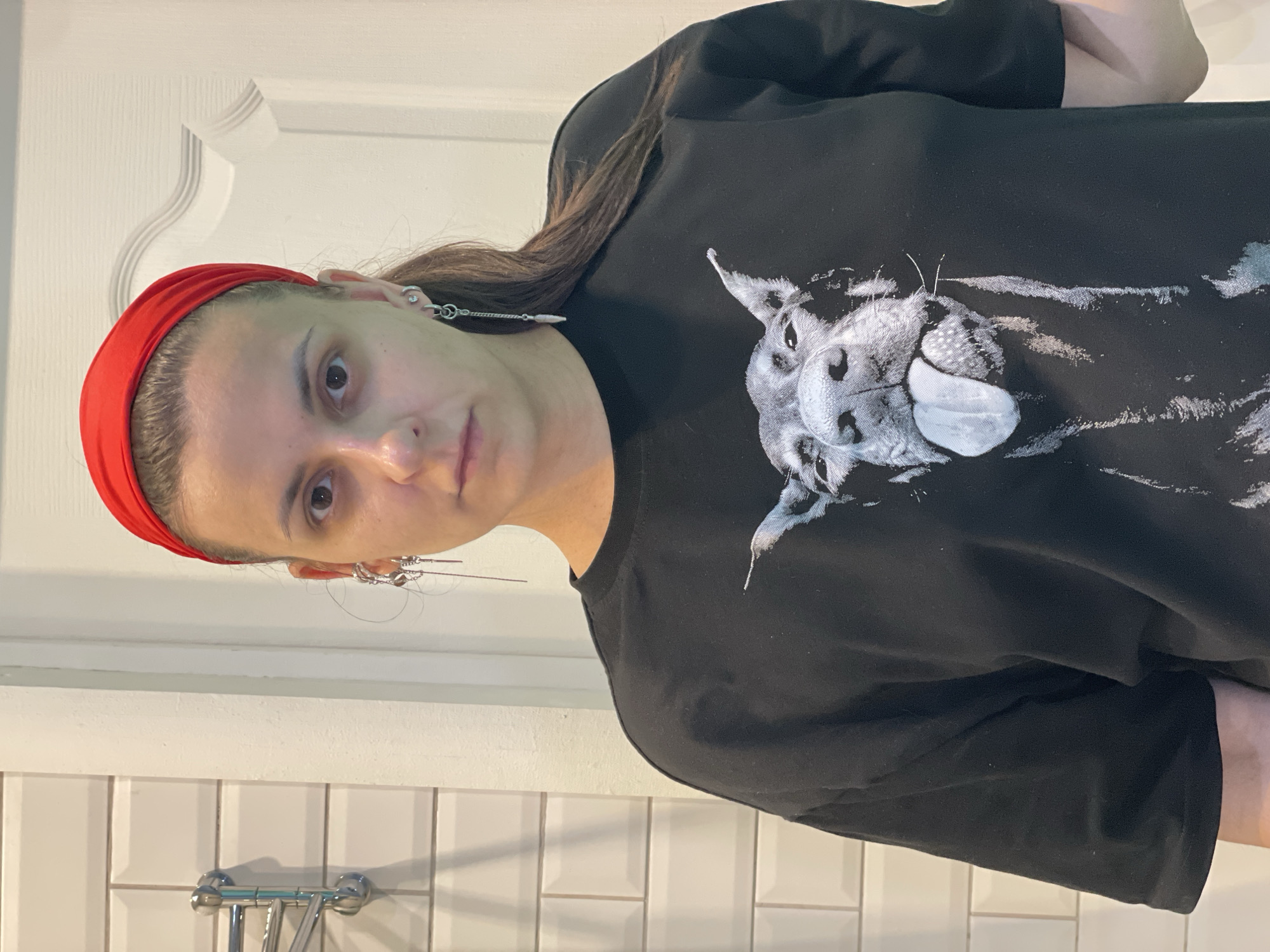 A female subject is depicted in a chest-up portrait, standing indoors and looking directly at the camera with a neutral expression. She wears a red fabric headband over her dark hair, which is pulled back. Both ears are adorned with multiple silver-toned piercings and dangle earrings; the subject's right ear (viewer's left) displays a long chain-style earring with a conical charm, while her left ear (viewer's right) features a long bar-style earring. She is clad in a black, short-sleeved garment, possibly a t-shirt or sweatshirt, featuring a prominent black-and-white graphic of a dog's head and upper body. The dog is depicted with its tongue extended, looking upwards. The background to the subject's left consists of a white tiled wall with dark grout, on which a chrome multi-bar fixture is mounted. To the subject's right, a white panel door with a decorative molded profile is visible. The scene is illuminated by even, interior lighting, located in the city of Unknown, Unknown.