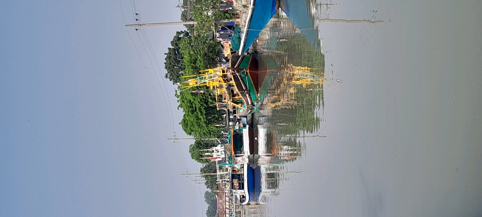 The image depicts a body of water with several boats moored along its edge. The boats appear to be fishing vessels, with prominent yellow structures and blue tarps visible. Lush green trees line the shore behind the boats, and utility poles with electrical wires are scattered throughout the scene. The water reflects the boats and the surrounding environment, creating a distorted but recognizable mirror image. The overall impression is that of a working harbor or waterfront in a tropical or subtropical region. This scene is likely located in the city of Pasuruan, Indonesia, a coastal city known for its fishing industry and port activities.