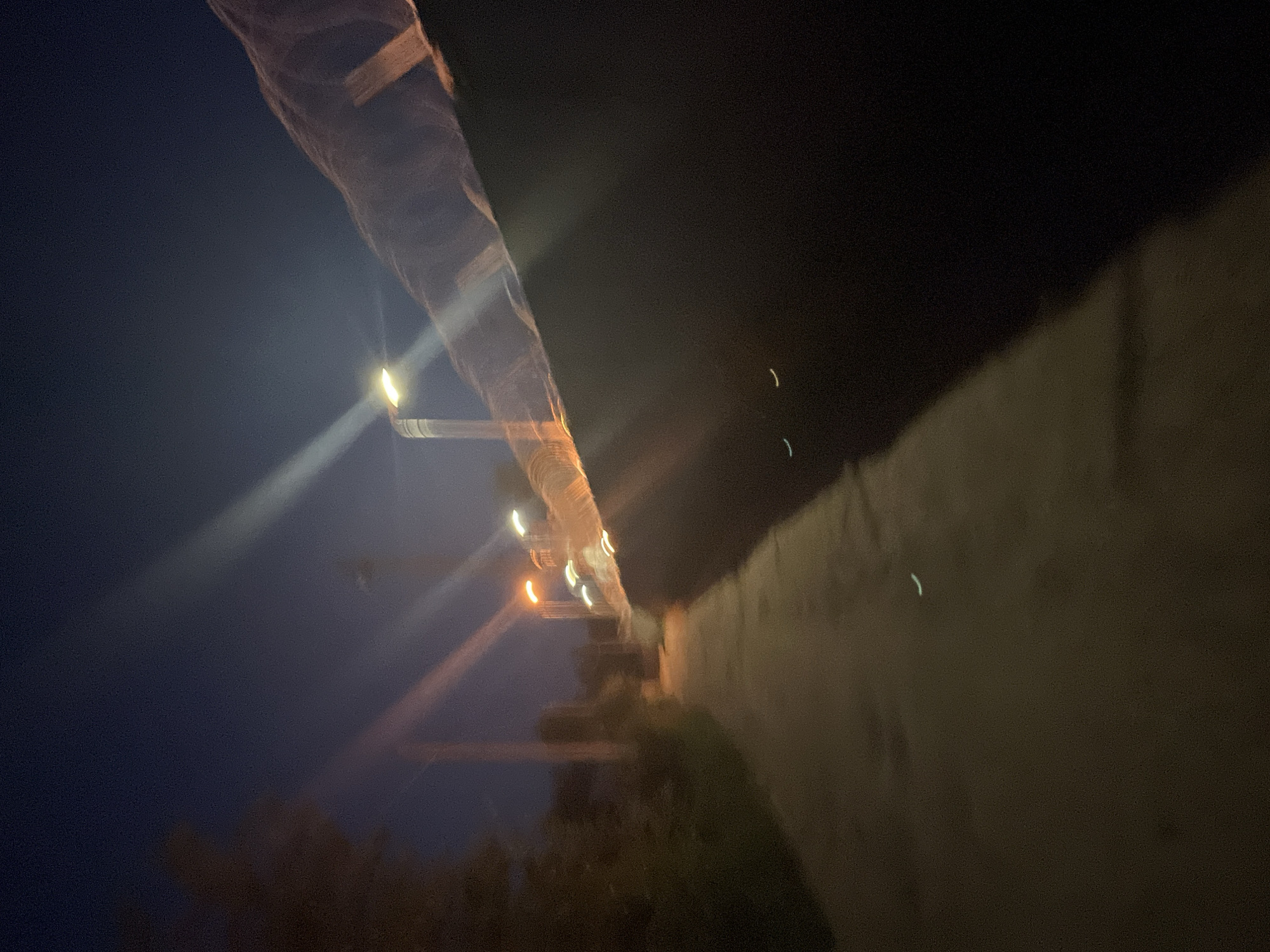 This is a night-time, slightly blurred photograph of a pathway next to a wall topped with barbed wire. The path appears to be made of asphalt or concrete and slopes slightly upwards into the distance. To the left of the path is an area with vegetation. Several streetlights are visible in the background, casting light streaks across the image. One streetlight is particularly prominent on the left, with a white light. Another streetlight, further down the path on the right, emits an orange glow. The sky is a deep blue, characteristic of twilight or night. The wall on the right is dark and imposing, with the sharp, metallic glint of barbed wire along its top edge. There are faint, curved light reflections on the dark surface of the wall, possibly from distant lights or the streetlights themselves. The overall impression is one of a somewhat desolate urban or industrial area at night, with the pathway leading towards the illuminated structures in the distance. Without additional context or visible landmarks, pinpointing the exact location within Chaikovsky, Russia is not possible based solely on the visual information in the image.