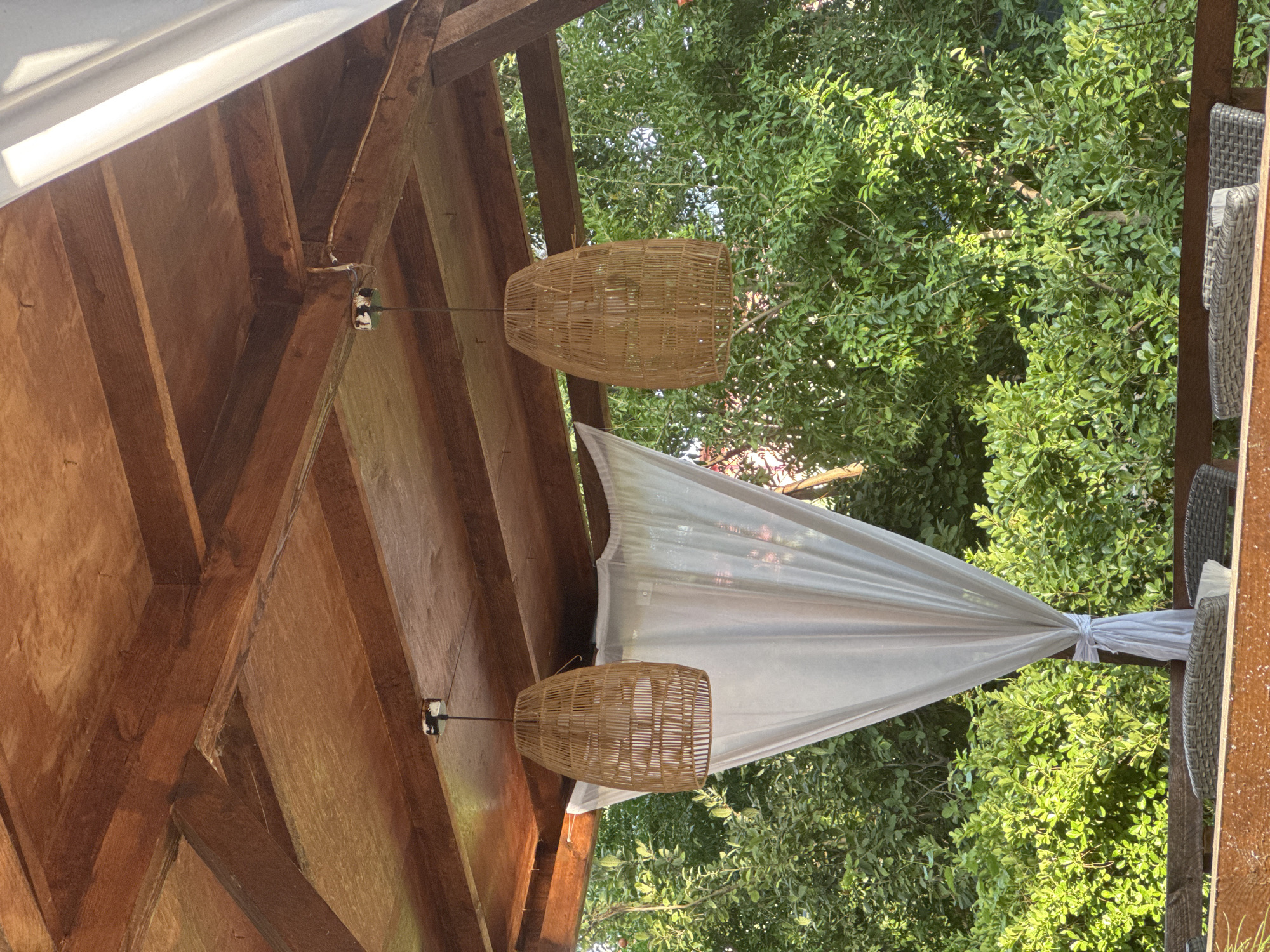 The image depicts an outdoor seating area, likely a cafe or restaurant, with a wooden roof structure. Two woven lampshades hang from the ceiling. A white sheer curtain is draped to the left, tied with a knot. Lush green trees and foliage are visible in the background. The scene appears to be a tranquil, natural setting. The specific location in Baku, Azerbaijan cannot be determined from the image alone.