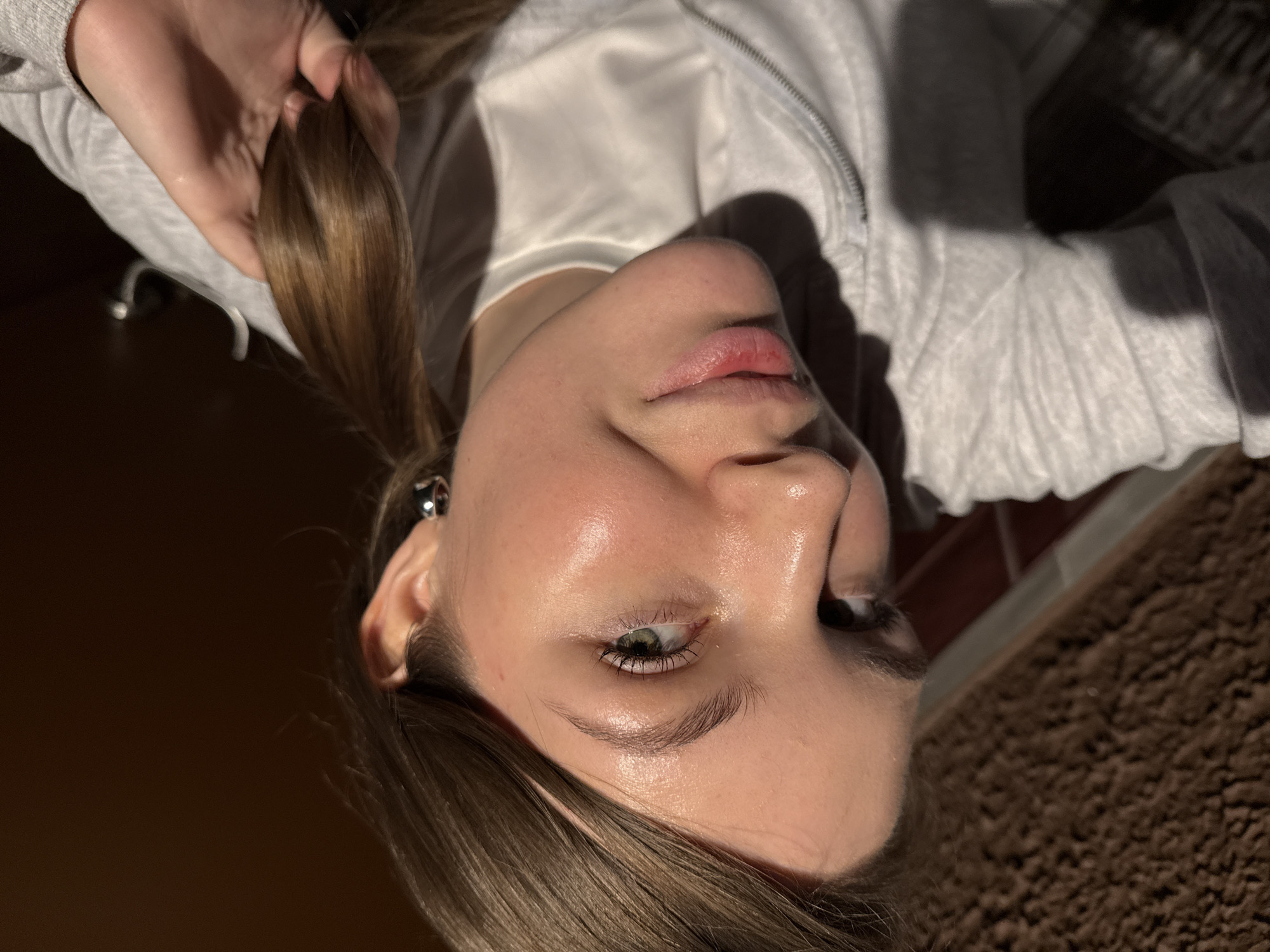 A close-up, tilted portrait shows a young woman with light skin, fair eyebrows, and green eyes. She is wearing a light-colored zippered sweatshirt. Her light brown hair is pulled back, and she holds a section of it in her hand. The background shows a blurred wooden floor, a carpet, and part of what appears to be a tiled wall. There is nothing to suggest the location is Belgrade, Serbia.
