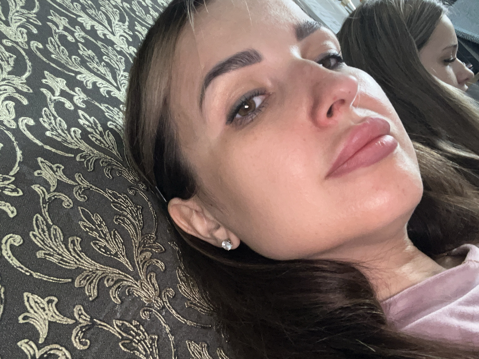 An extreme close-up shot shows a woman's face and shoulder against a dark, patterned wallpaper with a gold floral design. The woman's face is turned slightly to the right, with her left eye and eyebrow clearly visible. She has brown hair, large brown eyes, and full lips that are lightly colored. A diamond stud earring is visible in her left ear. She is wearing a light pink t-shirt. The overall lighting is bright, creating highlights on her skin. A reflection of the woman, also looking downwards, is visible on the right side of the image.