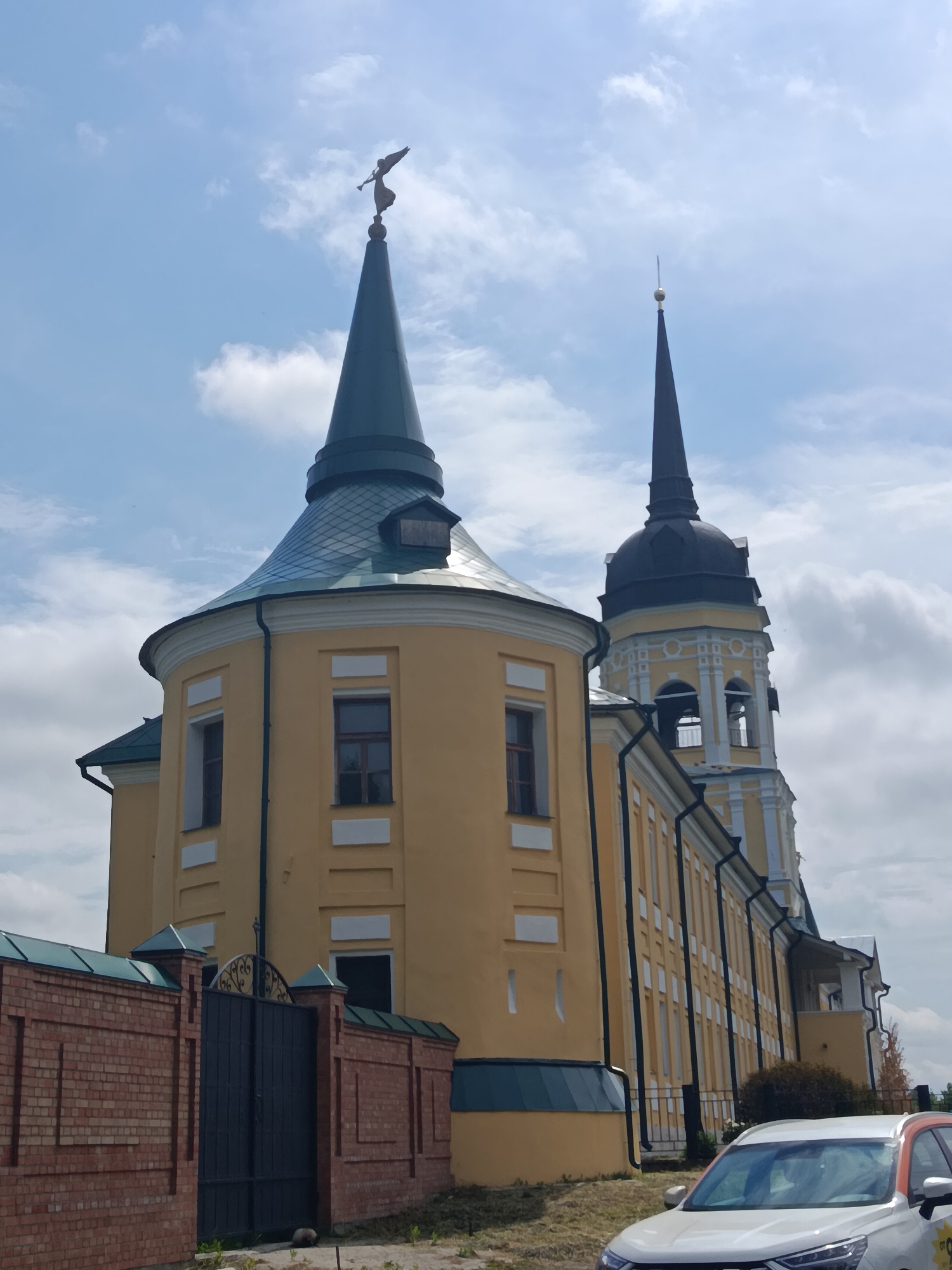 In Radovitsy, Russia, a tall, yellow, multi-story building with a distinctive conical roof is pictured. The building features multiple windows and a bell tower. A brick wall with a metal gate runs along the foreground, and a white car with orange accents is parked in front of the building. The sky is partly cloudy.