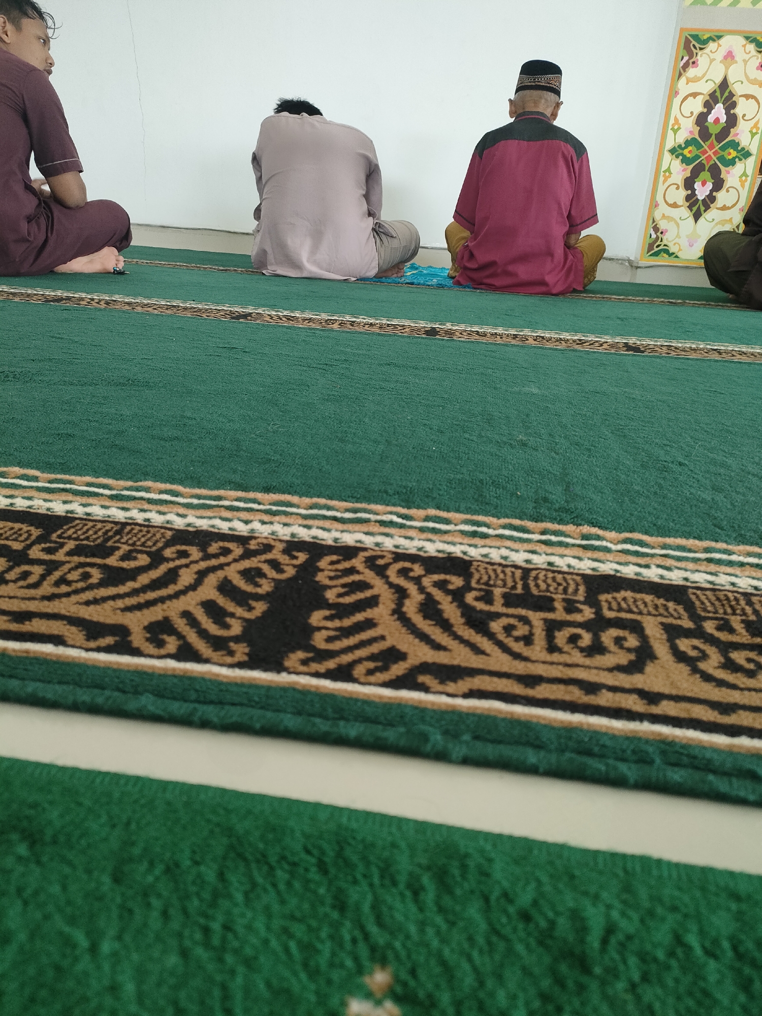 In Pringsewu, Indonesia, a group of men are sitting on a green carpeted floor in what appears to be a prayer room. Three men are visible from the back, and one from the side. The man on the far left is wearing a maroon outfit and is sitting with his legs crossed. To his right, another man is wearing a light purple shirt and beige pants, also seated. Further to the right, an elderly man in a maroon shirt with a black collar and a black and gold cap is seated in a cross-legged position. The man on the far right, partially visible, is wearing a dark green garment. The floor is covered with a green carpet that has a decorative border featuring intricate brown and black patterns. In the background, a white wall is present, with a colorful, ornate panel mounted on it, adorned with floral motifs in shades of green, pink, yellow, and brown. The lighting suggests an indoor environment, possibly during daytime.