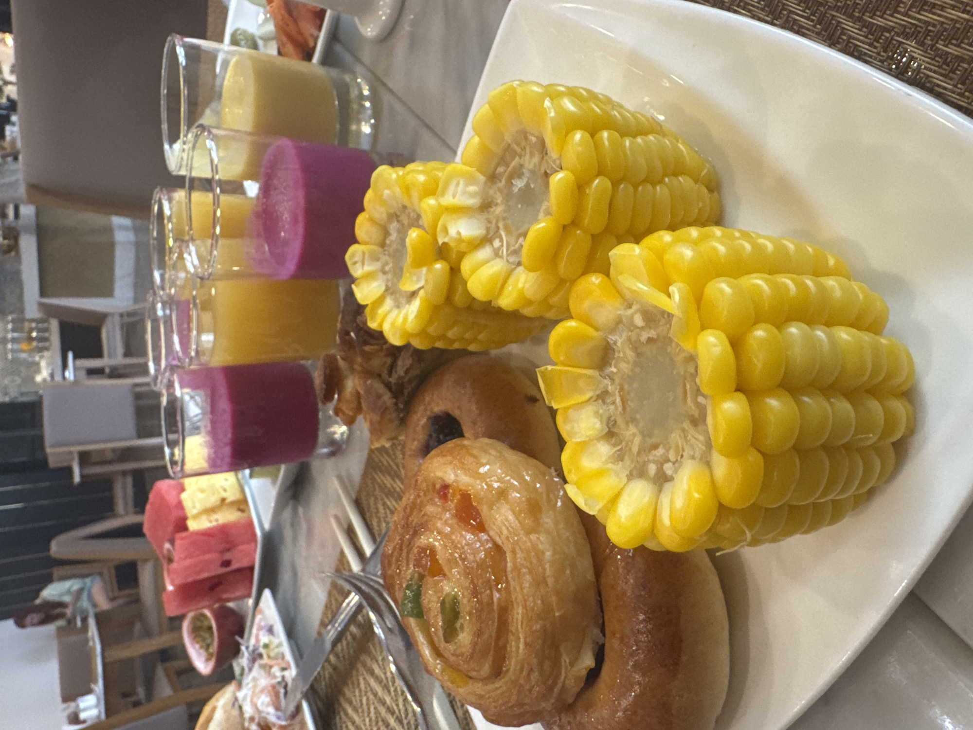 This close-up, eye-level shot features a plate of food, likely part of a buffet, possibly located in Đà Nẵng, Vietnam. The plate, which is white and curved, holds two pieces of cut corn on the cob, several small pastries, and doughnuts. There is a selection of fruit juices in glass containers to the left of the plate. A silver fork rests on the plate near the pastries. The food is displayed on a surface, possibly a table covered with woven material. In the background, there is a blurred view of a restaurant or dining area.