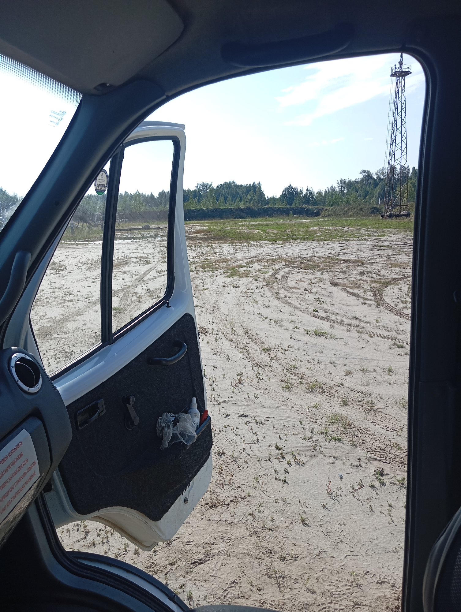 The image is taken from the interior of a vehicle, looking out the open driver's side door. The vehicle appears to be parked on a sandy, unpaved area with sparse vegetation. Tire tracks are visible in the sand. In the distance, a tall, lattice-like structure, possibly a transmission tower or a watchtower, stands against a clear blue sky with scattered white clouds. A line of trees forms a backdrop behind the open field. The interior of the vehicle shows part of the dashboard and the driver's side door panel, which has a small compartment containing a plastic bag with debris and a spray bottle. A label with text in Cyrillic is visible on the dashboard. The overall scene suggests an outdoor, possibly remote, location.