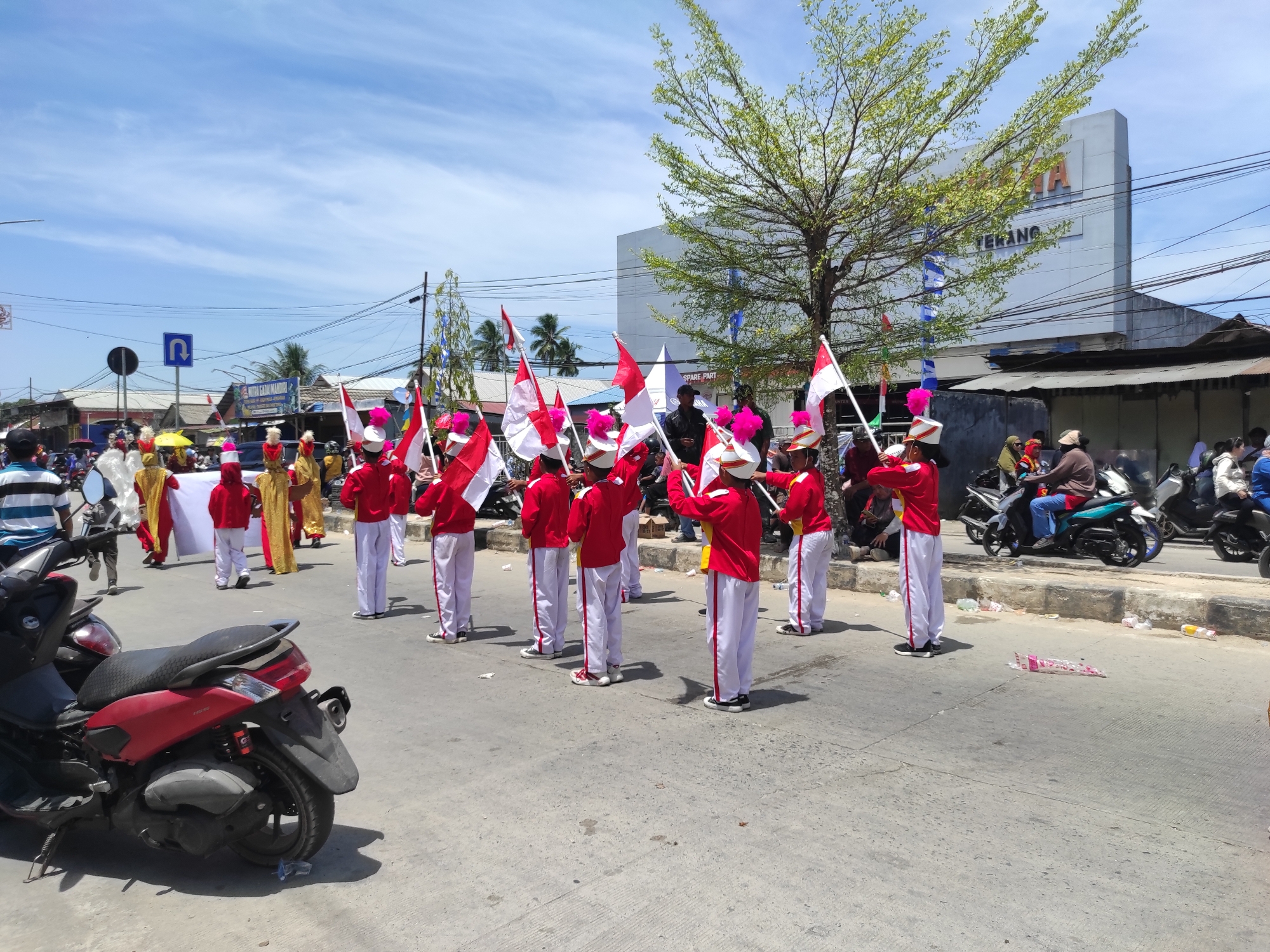 In Muara Jawa Ulu, Indonesia, on a bright, sunny day with a blue sky and scattered clouds, a group of individuals in marching band uniforms are participating in a parade. The participants, predominantly young, are dressed in red jackets with white sleeves and white pants, accented with red and white trim. Many are wearing white hats with pink plumes. They are holding and waving red and white flags, suggesting a patriotic or celebratory event. The parade is taking place on a paved street, with a red scooter parked in the foreground on the left. In the background, several buildings are visible, including a large white structure with signage that appears to be a commercial establishment. Power lines and trees are also present. Other individuals and vehicles, including motorcycles, are present along the sides of the street, observing the parade. The environment appears to be an urban or semi-urban setting.
