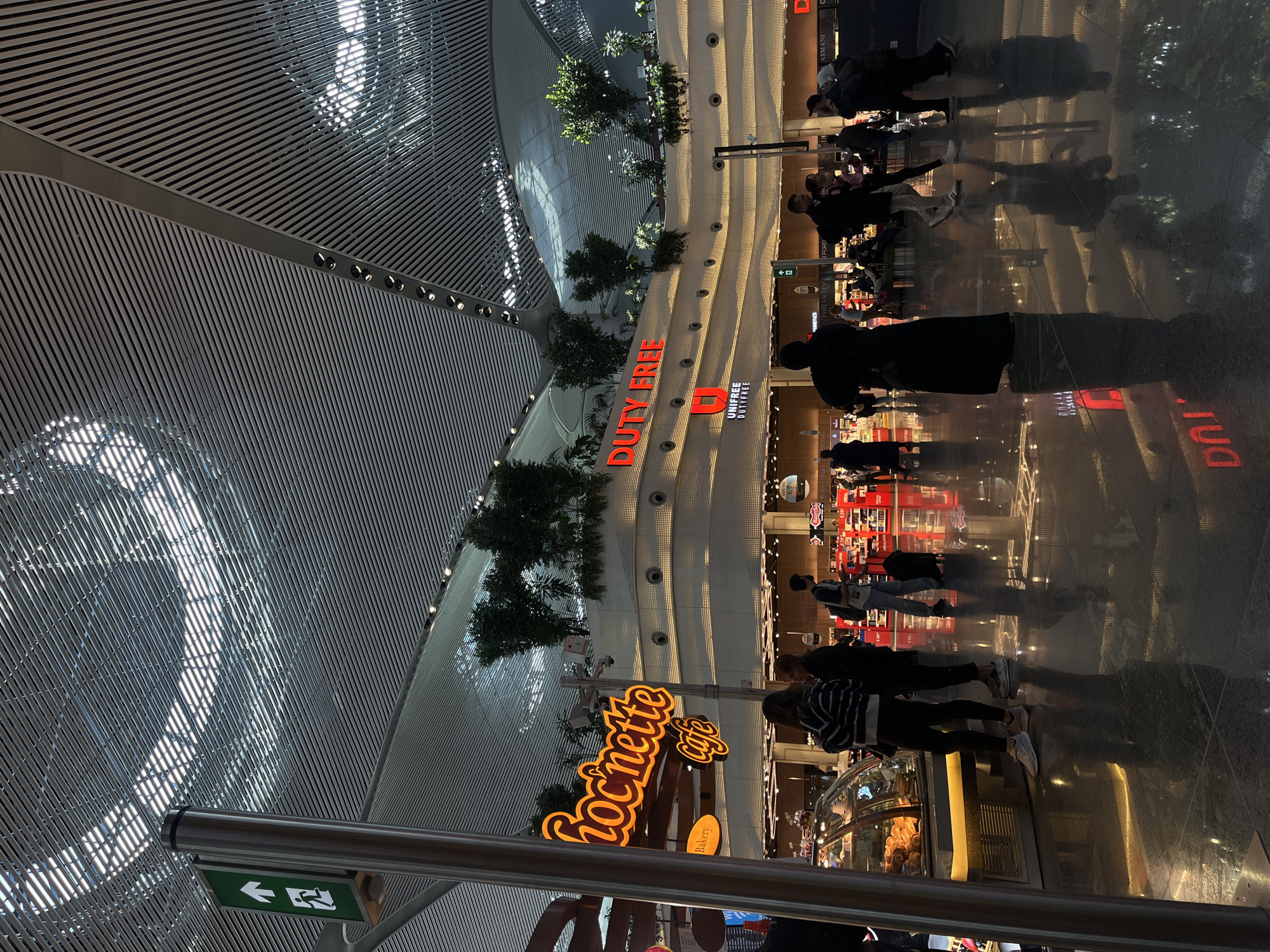 The image depicts the interior of a modern airport terminal, specifically Istanbul Airport in Istanbul, Turkey. The scene is illuminated by artificial light and displays a multi-level commercial area.
The environment features a high, curved ceiling composed of numerous parallel, light-colored metal slats, with integrated linear lighting elements and scattered brighter sections. The polished dark tiled floor exhibits strong reflections of the overhead lighting and illuminated signs. A prominent architectural structure forms the mid-ground, featuring curved tiers with horizontal bands of small, recessed circular lights and integrated planters containing green foliage and small trees on its upper levels.
In the foreground to the left, a silver metallic pole stands vertically, fitted with a green sign displaying a white upward-pointing arrow and a white pictogram of a person running, indicating an exit or direction. To the left-midground, an orange neon sign reads "Chocnette cafe" in a cursive font, with a smaller "Bakery" sign below it. A glass display counter with pastries is positioned beneath these signs. To the right-midground, a large red neon sign prominently displays "DUTY FREE," with a smaller "UNIFREE DUTYFREE" logo below it.
Approximately 15 to 20 individuals are visible throughout the scene, primarily in the mid-ground and background. They are casually dressed and appear to be travelers or shoppers. Two females stand near the "Chocnette Cafe" display counter, one wearing a striped long-sleeved shirt and dark pants, the other a dark jacket and dark pants. Multiple individuals are walking across the polished floor, some with luggage. A male in a light shirt is seen pulling a black roller suitcase. Another individual in a long dark garment stands with their back to the camera in the center-right. The reflections on the floor clearly show the lower half of many individuals and the inverted red "DUTY FREE" sign. Actions observed include walking, standing, and observing merchandise displays. No specific interactions between individuals are clearly discernible.