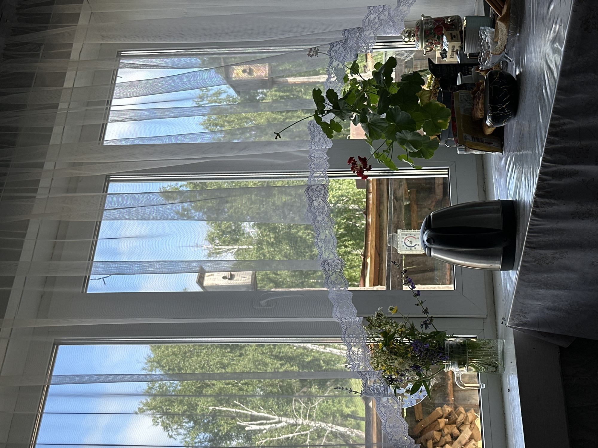 The image depicts an indoor scene looking out a window. The window is divided into three panes and covered by sheer white curtains with a decorative lace trim. Outside, lush green trees and a bright blue sky are visible. Two wooden birdhouses are mounted on poles in the distance. On the windowsill, a glass jar filled with a bouquet of wildflowers sits to the left. Next to it is a stainless steel electric kettle, and further to the right, a potted green plant with red flowers is displayed. Behind the plant, various items are arranged, including boxes, a decorative teapot, and a small bowl containing what appears to be sugar. A table covered with a patterned tablecloth is in the foreground, with a black and white patterned bowl and some bread on it. The overall impression is a view from inside a rustic home, likely in a rural or suburban setting given the presence of trees and birdhouses. There are no people visible in the image.