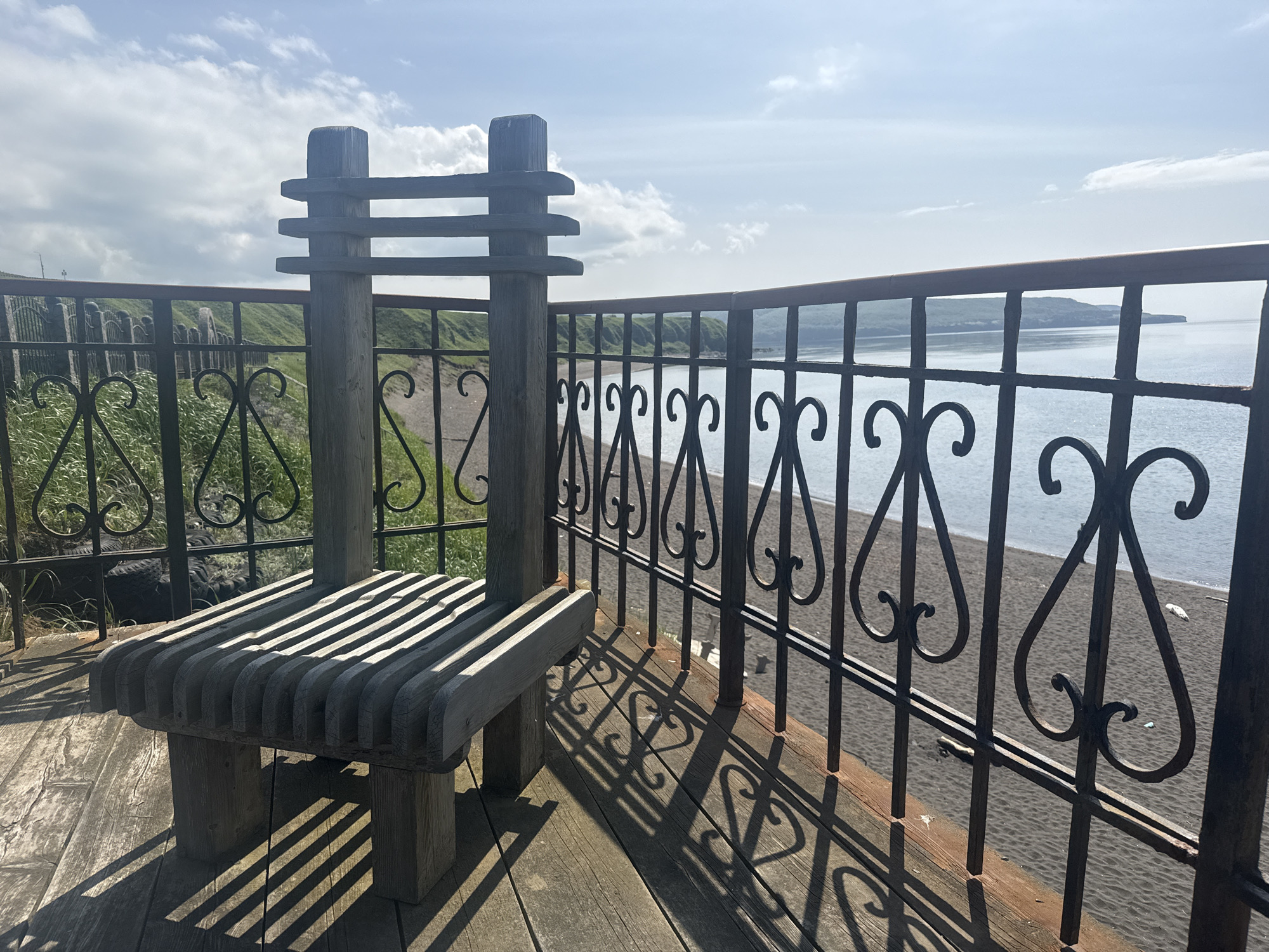 A rustic wooden chair and bench sits on a wooden deck overlooking a beach and the sea. The chair and bench are made of rough-hewn wood, with a slatted seat and back. A black metal railing with decorative scrollwork separates the deck from the beach. The beach is dark sand, with calm, light blue water. In the background, a green, rolling hillside slopes down to the sea. The sky is blue with scattered white clouds. This scene appears to be in a coastal town.