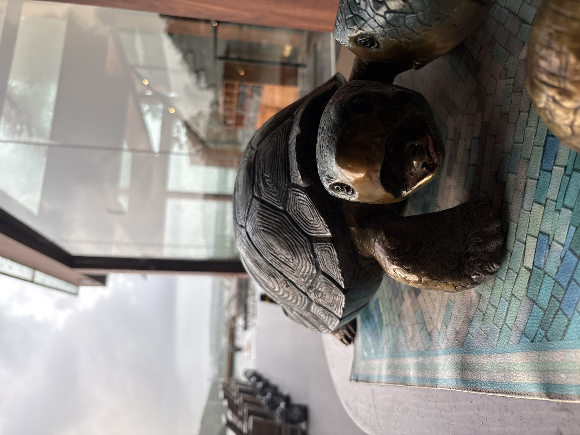 A close-up, low-angle shot shows a bronze turtle sculpture with an opening in its mouth, resting on a patterned mat. The turtle is positioned on a table, with another turtle sculpture partially visible to its right. In the background, a row of empty chairs and a blurred view of a body of water with a bridge are visible, suggesting an outdoor seating area. The scene is set against a backdrop of a large glass window reflecting the sky and interior elements of a building. The image appears to be taken in Vagator, India, possibly at a restaurant or resort.