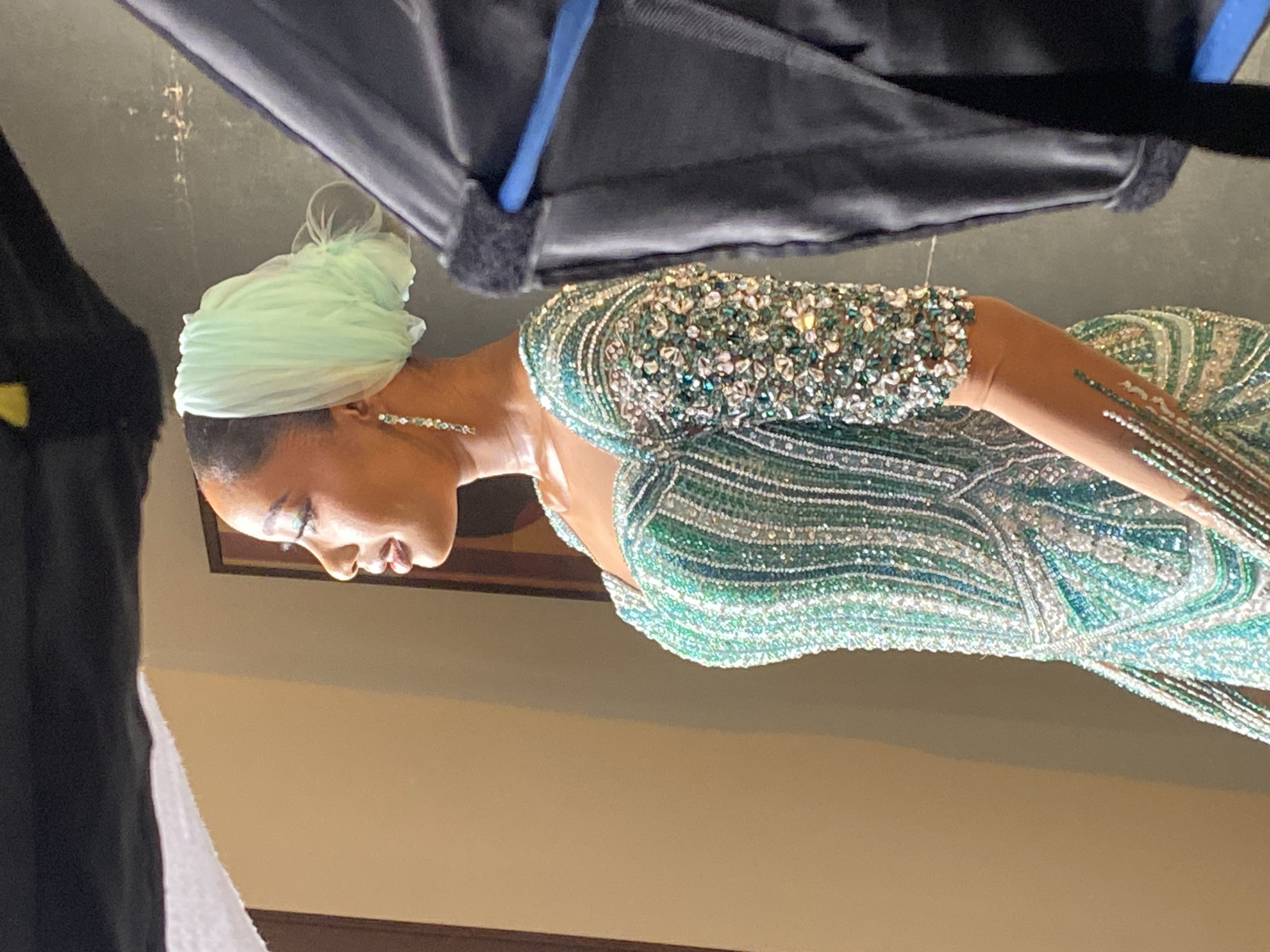 A woman is wearing a light green head wrap and a green beaded gown. The background appears to be an interior space. The location is Unguwar Sarki, Nigeria.