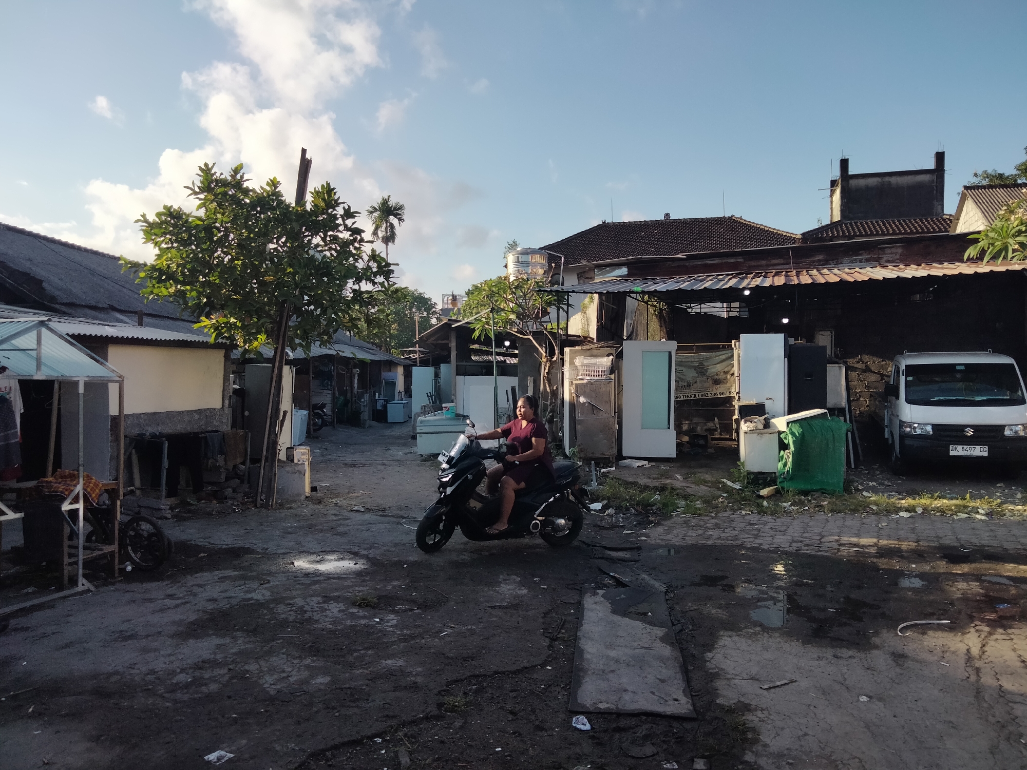 A woman rides a black motorcycle on a dirt road in Denpasar, Indonesia. She is wearing a burgundy dress and has dark hair. The road is wet and has puddles. There are several buildings and structures on either side of the road, including a white van parked on the right. The sky is blue with some clouds. The overall scene appears to be in a semi-urban or rural area, possibly a back alley or a less developed part of the city. The environment is somewhat cluttered with various objects, suggesting a working or storage area. The lighting indicates it is likely late afternoon or early morning.