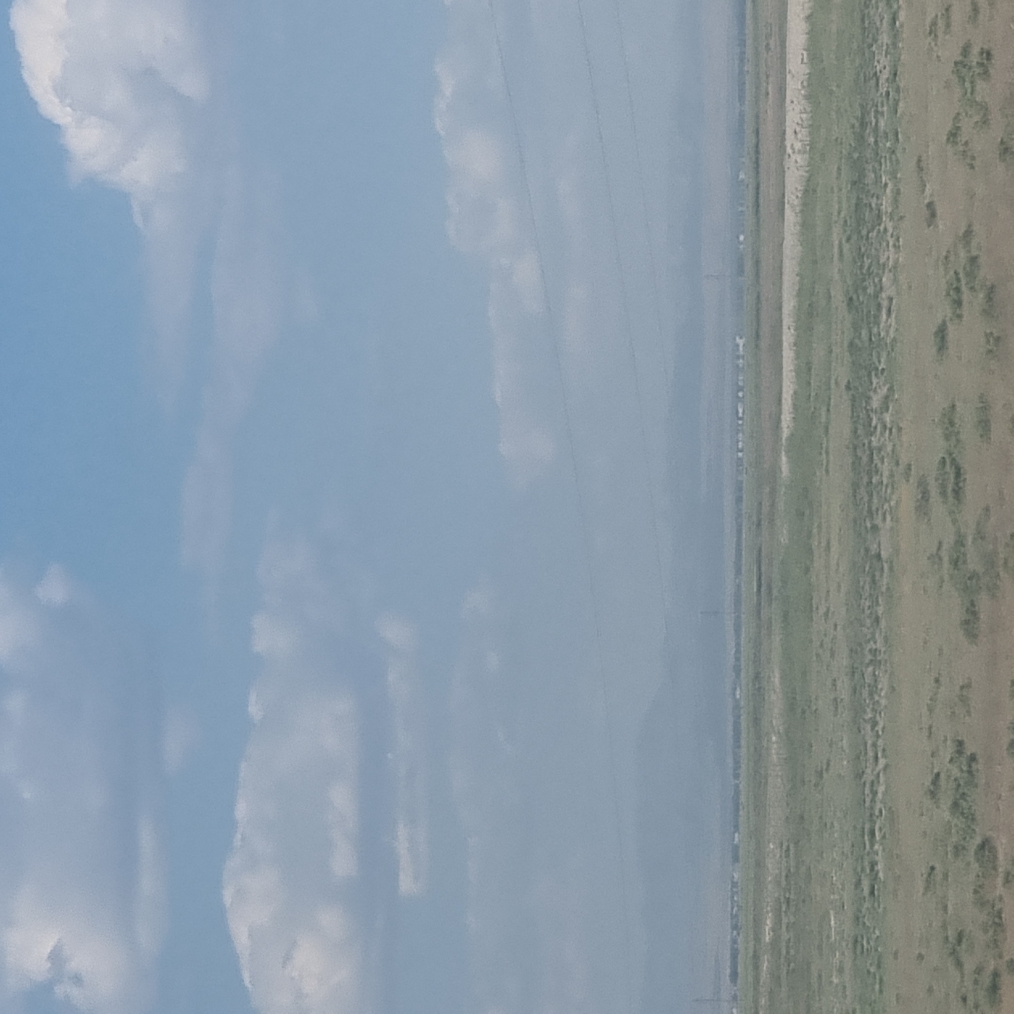 The image depicts a wide, flat landscape under a hazy sky with scattered clouds. The foreground consists of dry, sparse vegetation, appearing as patches of brown and green grass. In the middle ground, a band of lighter-colored earth runs horizontally across the scene, separating different areas of vegetation. Further in the distance, a faint, blurry line of structures suggests a settlement or town. A series of power lines are visible as thin dark lines stretching across the sky. The background is dominated by a hazy mountain range, almost lost in the atmospheric conditions. The overall impression is one of a dry, open expanse, possibly in a rural or semi-arid area. The location is identified as Shoytobe, Kazakhstan.