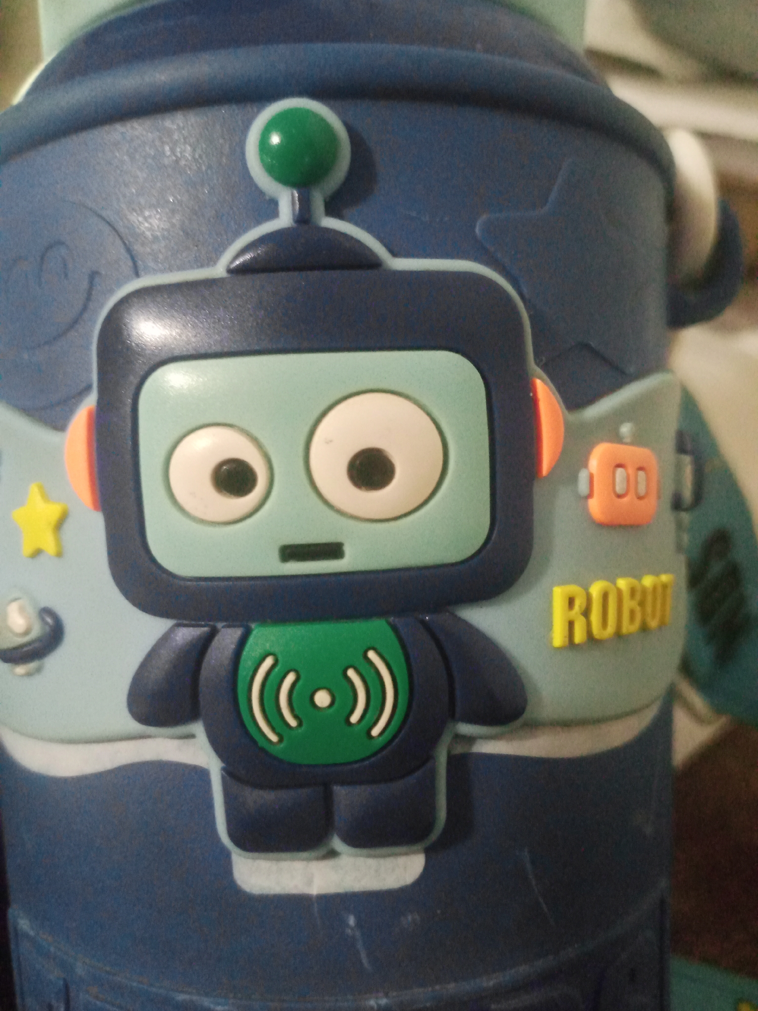 The image shows a close-up of a blue object, possibly a water bottle or a container, with a cartoonish robot decal. The robot has a light blue head with large white eyes, orange circular ears, and a dark blue body. On the robot's chest is a green circle with white concentric arcs and a central dot, suggesting a signal or sound wave. To the right of the robot, the word "ROBOT" is printed in yellow, stylized letters. There is also a small orange rectangular icon with two circles on it, resembling a simplified robot face. The background is a textured blue surface with some lighter blue elements, including a yellow star and a small grey object. The lighting is somewhat dim, and the image is slightly blurry, suggesting it was taken in low light conditions. There is no indication of Santa Ana, Venezuela, in the image.