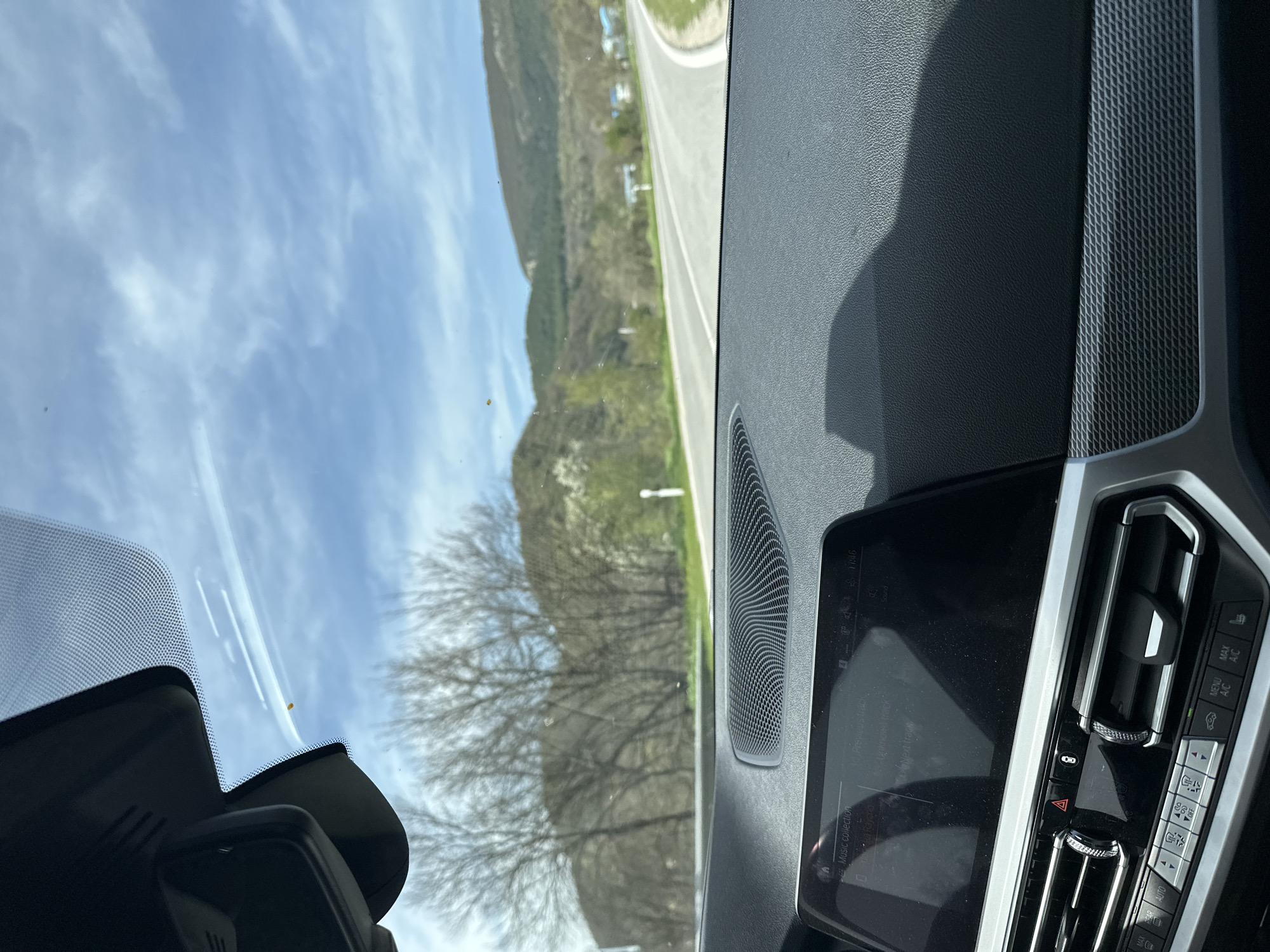 The image captures a view from inside a modern car, looking out through the windshield at a rural landscape. The perspective is from the passenger side, showcasing a significant portion of the car's dashboard, infotainment system, and the external scenery.
Inside the car, the dashboard features a textured, dark gray material, with a patterned trim panel running along the passenger side. A speaker grille is visible near the windshield. The infotainment system display shows "Music collection" and the time "11:46", along with "Previous track" indicating audio playback. Below the screen are climate control buttons and dials, including "AUTO", "MENU", "A/C", "MAX A/C", fan speed controls, temperature adjustments, and the hazard light button. The top of the windshield shows the housing for the rearview mirror or driver-assist cameras, with a dotted frit band along its edge.
Outside the vehicle, a clear blue sky with scattered white cirrus clouds dominates the upper portion of the view. A paved road, appearing to curve slightly to the right, stretches into the distance, flanked by green verges. Beyond the road, rolling hills or mountains are visible, covered in sparse, early spring vegetation and some bare-branched trees. The landscape suggests a journey through a scenic, perhaps mountainous, region. The bright sunlight and clear skies indicate a pleasant, sunny late morning.
The overall scene depicts a peaceful drive or stop in a natural setting. The visible text on the infotainment screen includes "Music collection", "11:46", "Previous track", and controls like "AUTO", "MENU", "A/C", "MAX A/C", and "OFF". The location context indicates this scene is set in Madrets, Bulgaria.