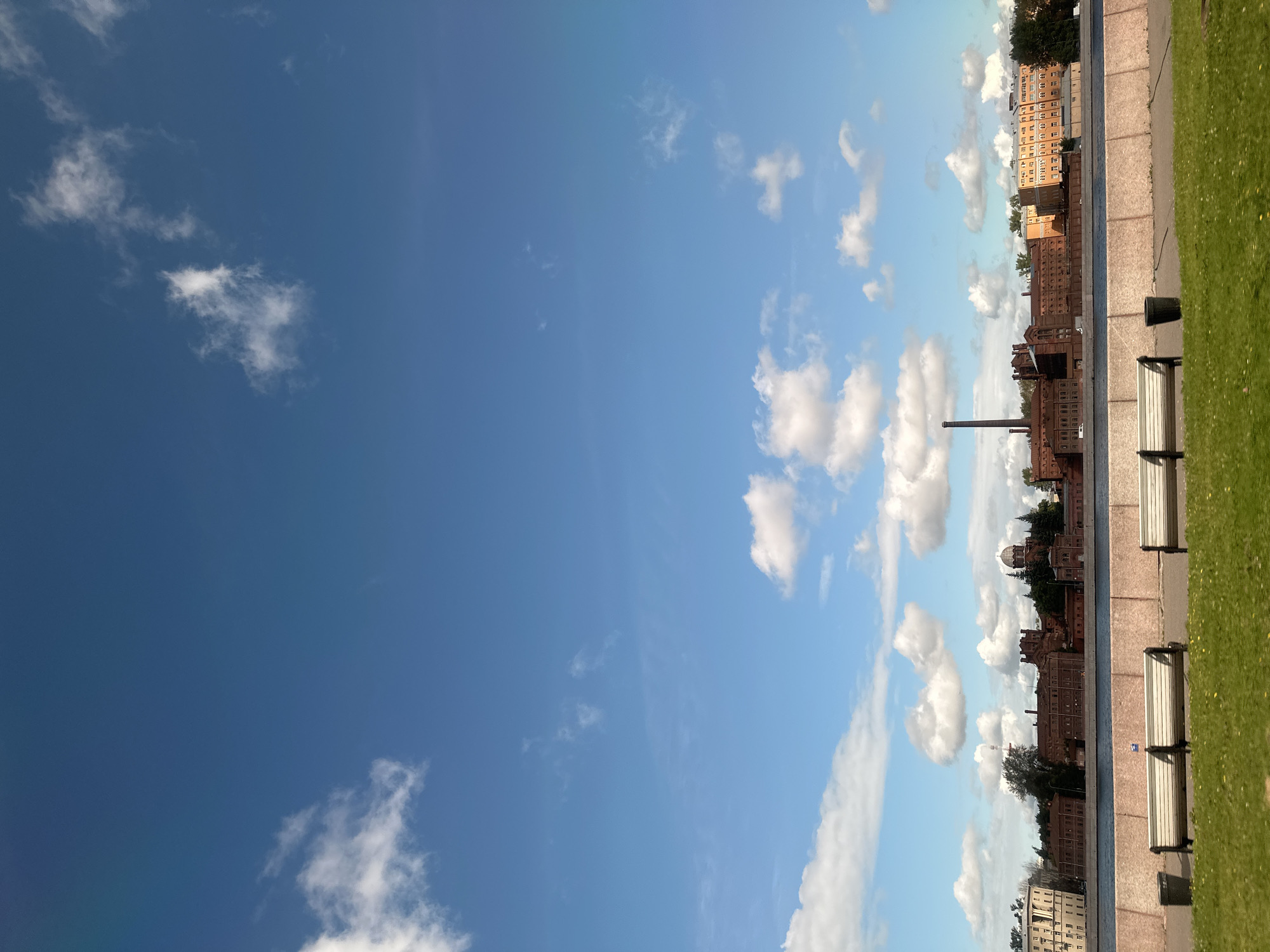 The image depicts a sunny day in Saint Petersburg, Russia, with a wide expanse of blue sky dotted with white clouds. In the foreground, a grassy area with scattered yellow flowers borders a paved walkway. Along the walkway are three park benches and two trash cans. A concrete barrier separates the walkway from a body of water, likely a canal or river. Across the water, a long, historic brick building complex stretches along the embankment. This complex features numerous windows, multiple sections of varying heights and architectural styles, and a prominent smokestack. Further down the embankment, lighter-colored modern buildings are visible, interspersed with trees. The architecture of the brick complex, with its crenellated towers and industrial appearance, suggests it may be a former factory or prison. The presence of such historic industrial buildings along the waterways is characteristic of Saint Petersburg's industrial heritage. The scene appears calm and tranquil, with no people visible in the frame.