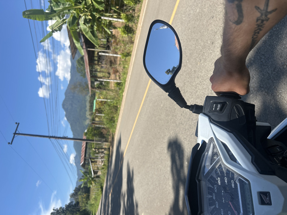 The image depicts a first-person perspective from a white Honda VARIO scooter on a rural road in Ban Mai Saha Sampan, Thailand, under a clear blue sky. The right hand of an individual, displaying forearm tattoos, grips the scooter's throttle. The scooter's dashboard shows a speedometer marked up to 160 km/h and an "IDLING STOP" button on the right handlebar control cluster. The right side mirror reflects a portion of a traditional Thai structure with a pointed roof spire, trees, and blue sky.
The asphalt road features a solid yellow line. To the right of the road, rural structures with corrugated metal roofs are visible behind a fence of wooden posts and wire. Lush green vegetation, including banana plants, lines the roadside. A utility pole with numerous power lines stands prominently. In the background, dark green, forested mountains rise under a sky with scattered white clouds. Shadows from objects to the left indicate strong overhead sunlight.