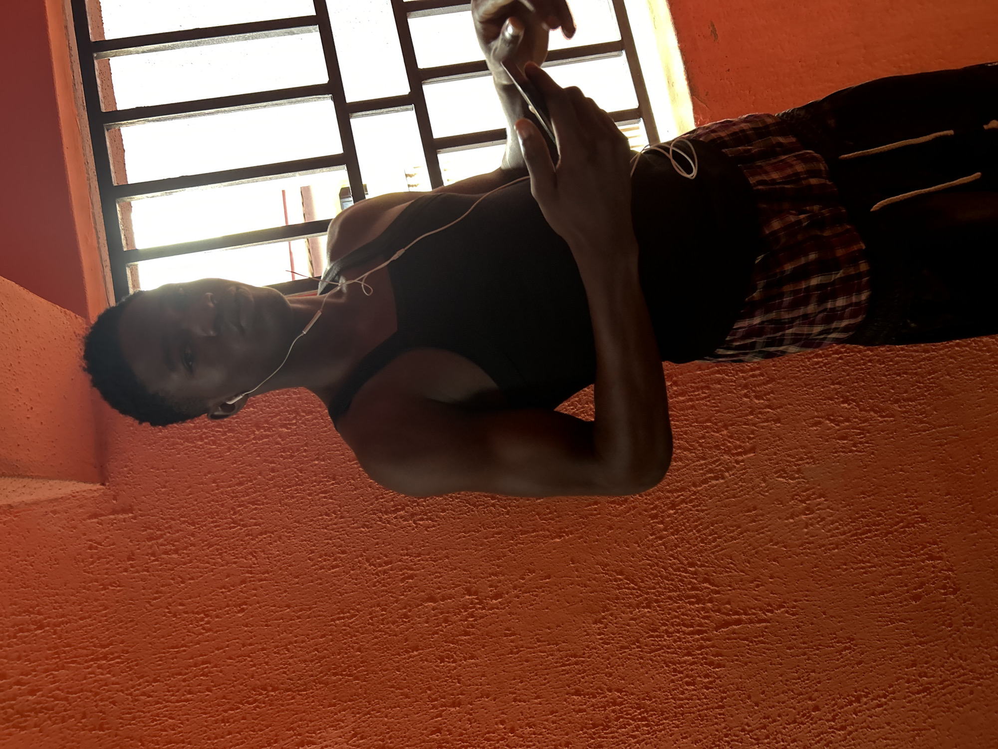 In the city of Ihiagwa, Nigeria, a young Black male stands against a textured orange wall. He is wearing a black tank top, red and white plaid shorts, and black sweatpants. White earbuds are in his ears, with the cord trailing down. He is holding a black smartphone in his right hand, his left hand extended towards it. Behind him, a barred window allows bright light to enter, casting shadows and silhouetting part of his body. The scene appears to be indoors, possibly in a residential setting.