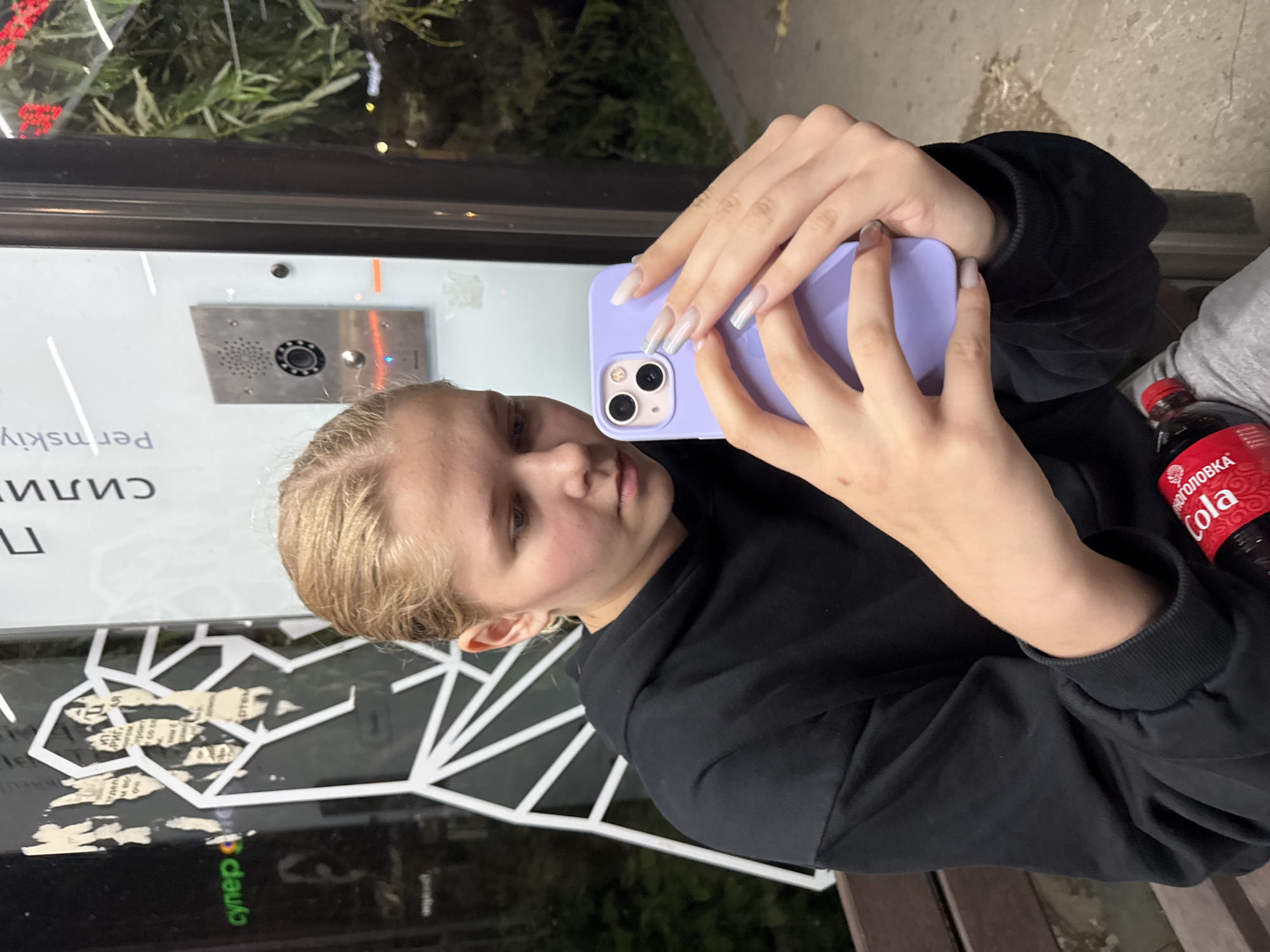 A young woman with blonde hair, wearing a black hoodie and gray sweatpants, is sitting outdoors and taking a selfie with her purple smartphone. She has long, acrylic fingernails. A bottle of Coca-Cola is visible next to her. Behind her is a glass window with graphics, including an abstract white geometric design and Cyrillic text. To the right of the window, there is a metal intercom panel. The background shows greenery and some scattered debris on the ground, suggesting an urban environment in Perm, Russia, possibly near an entrance to a building.