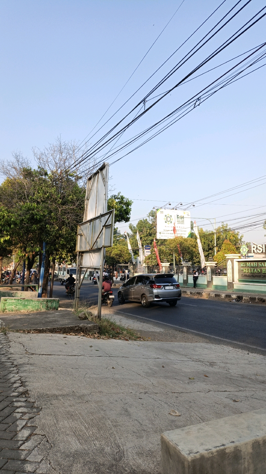 The image shows a street scene in Kuwasen, Indonesia. A silver car is turning right on a road with two lanes. In the background, a hospital building with the sign "RUMAH SAKIT 'SULTAN HADI'" is visible. Several trees line the road, and there are power lines overhead. A billboard is also present on the left side of the image. People on motorcycles are seen on the road and sidewalk. The weather appears to be clear and sunny.