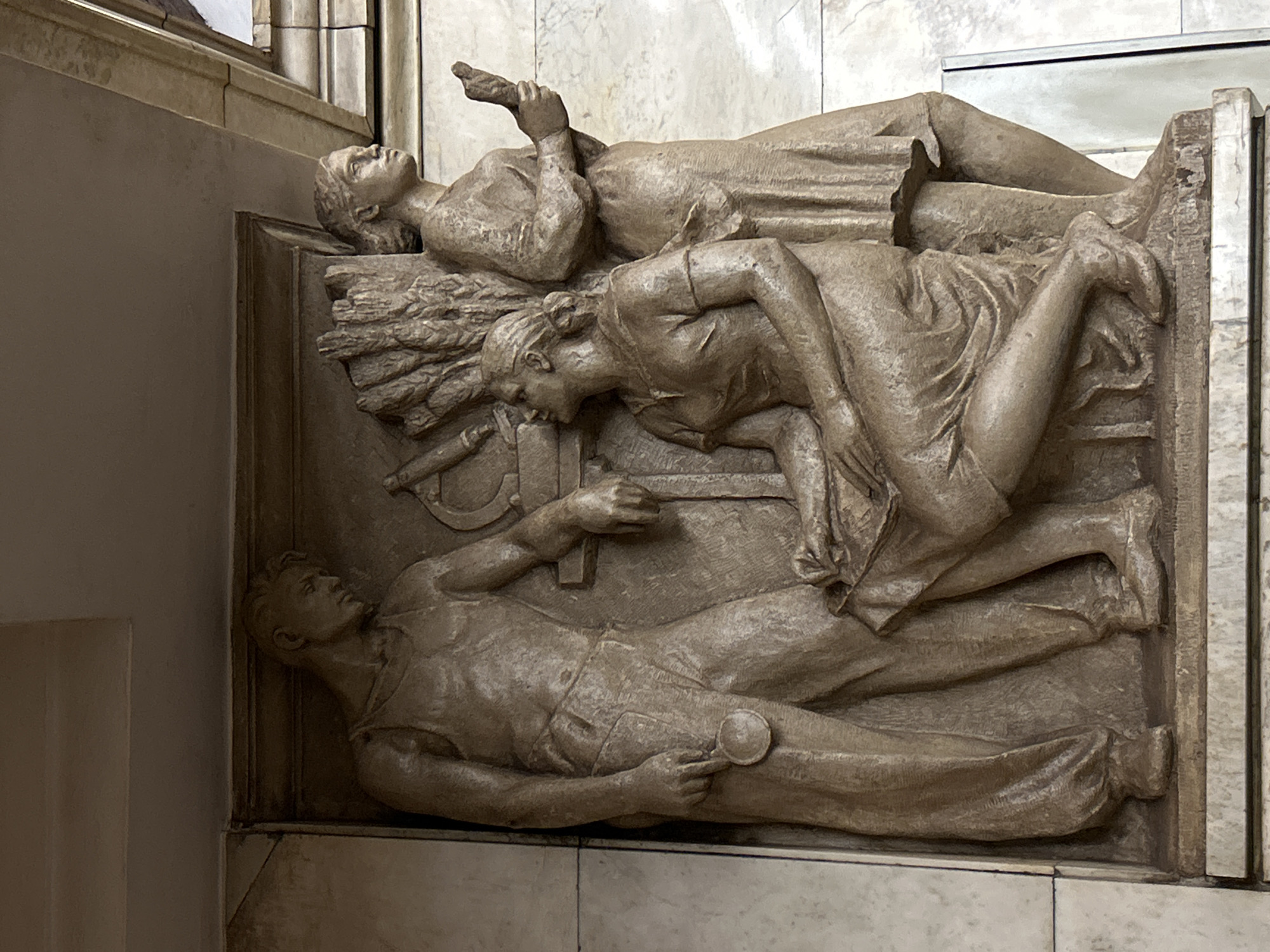 The image depicts a bas-relief sculpture located in Saint Petersburg, Russia. The sculpture features three figures: a man, a woman, and another individual whose gender is less discernible. The man lies at the bottom, holding a magnifying glass. Above him, the woman leans forward. The top figure is reclined with a raised hand holding an object. They are surrounded by symbols of labor, including a bundle of wheat and tools such as a hammer and sickle. The sculpture is mounted on a wall with marble-like surfaces.