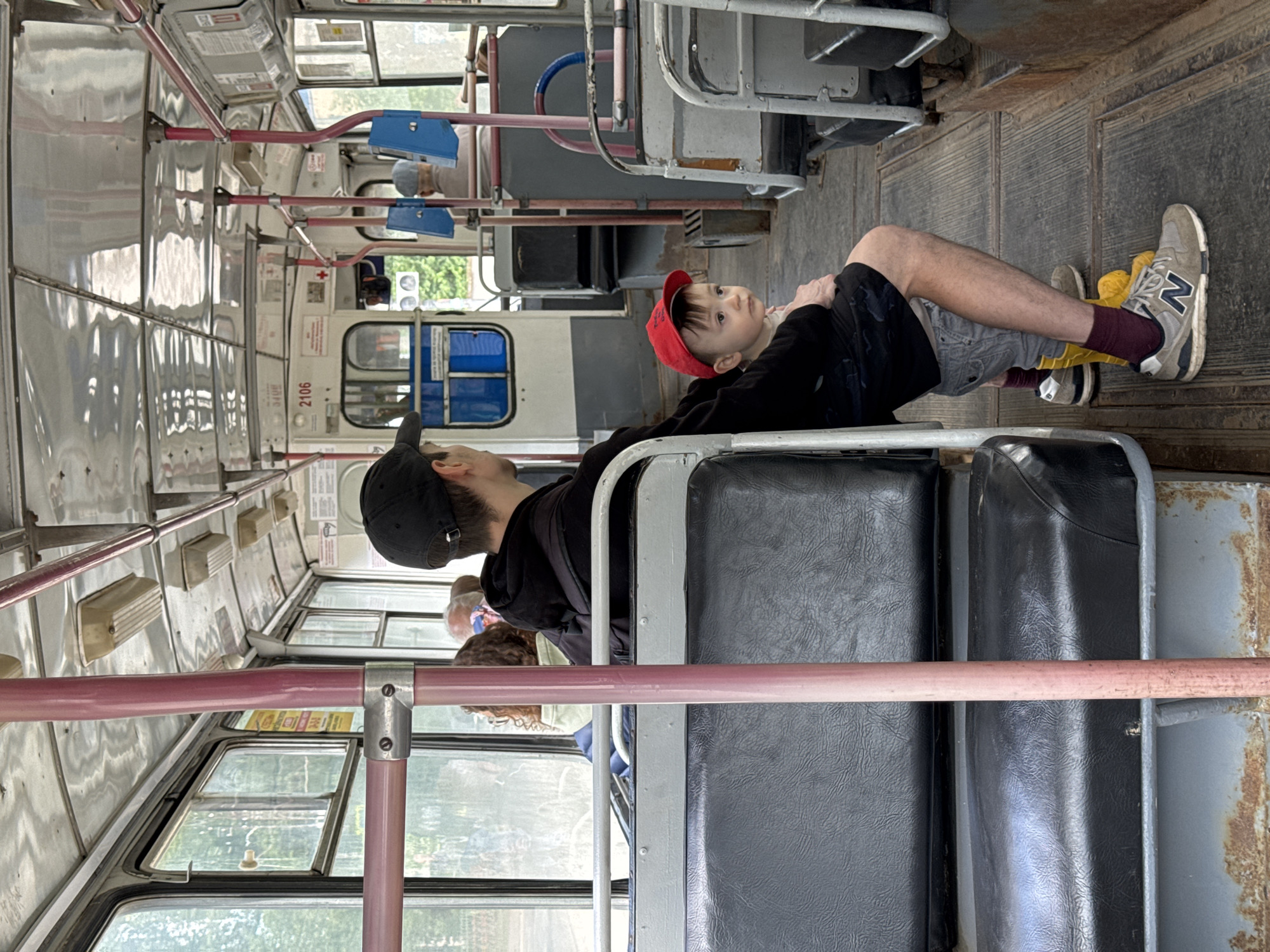 The image shows the interior of a trolleybus, likely in the city of Izhevsk, Russia, based on the Cyrillic lettering and appearance of the bus. Two people are visible. An adult wearing a black cap and a black jacket sits on a seat, facing forward. A child wearing a red cap is seated adjacent to the adult, with their legs stretched out, feet resting on a seat. The interior of the bus is weathered with visible signs of wear and tear. Other passengers are visible in the background. A pink horizontal handrail is in the foreground.