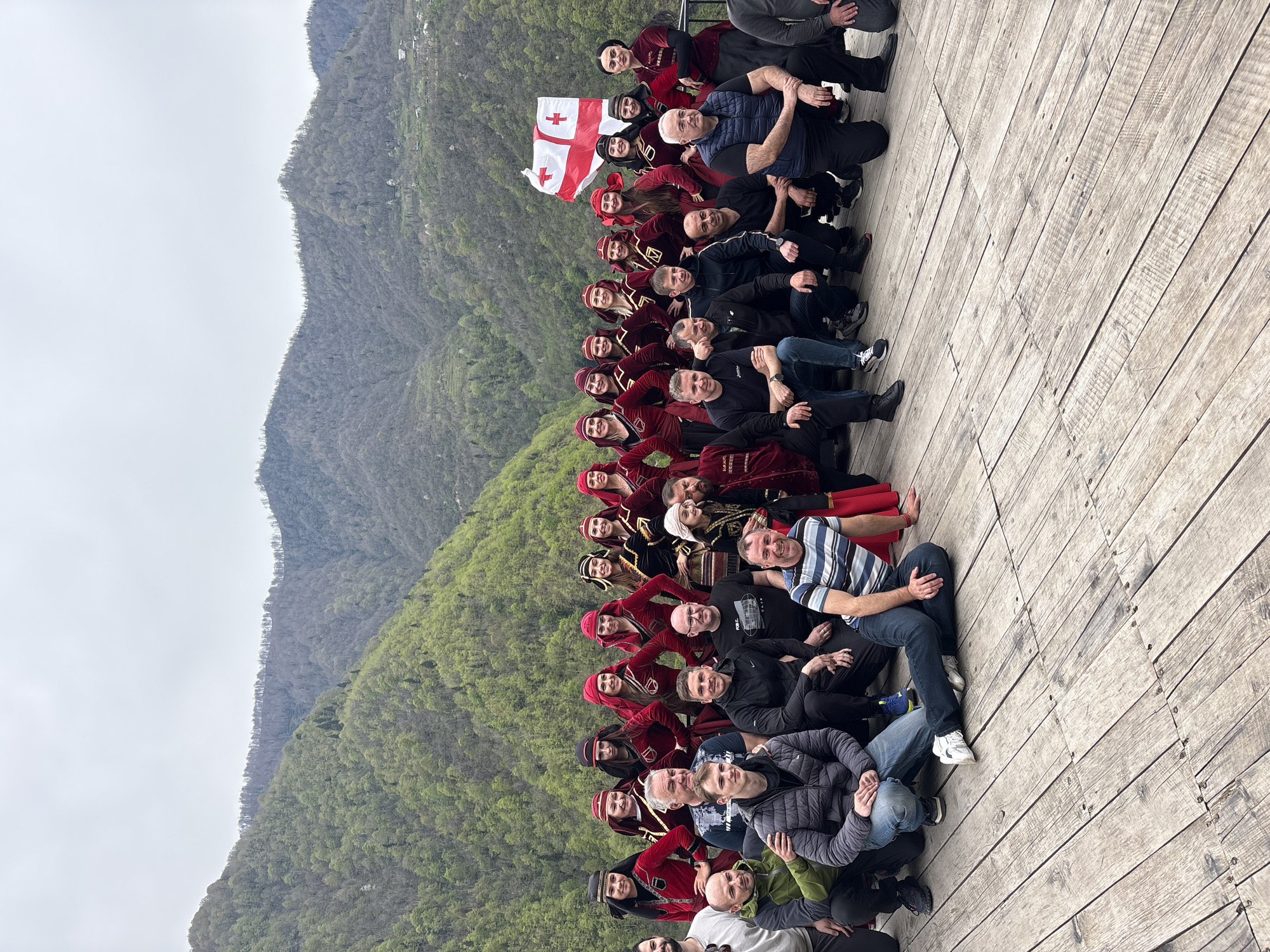 A group of approximately 40 people are seated on a wooden platform, posing for a photo. The majority of the group are wearing traditional Georgian clothing, specifically red velvet chokhas and hats. A Georgian flag is visible in the middle of the group. The platform overlooks a mountainside with a wooded area. The photograph was taken in Kveda Chkhutuneti, Georgia. The sky is overcast.