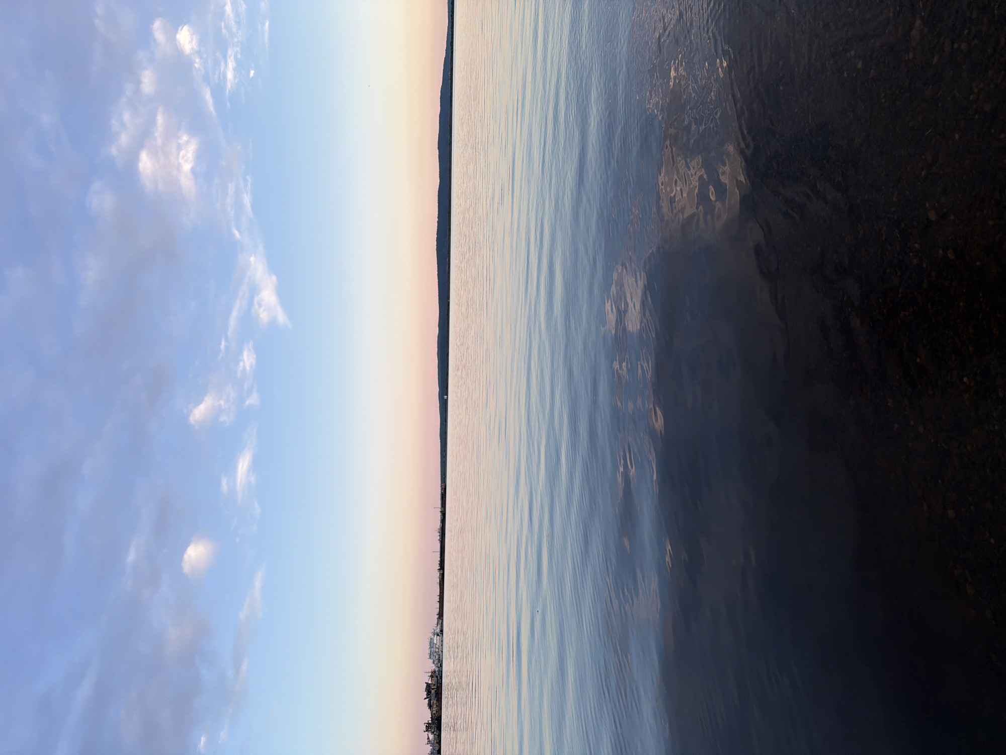 The image displays a serene landscape featuring a wide body of water beneath a partially cloudy sky. The composition is horizontally bisected by a distant horizon line, beyond which a faint, dark strip of land is visible.
The upper portion of the image is dominated by a sky that transitions from a clear, pale blue near the horizon to a deeper blue overhead. Scattered white and light gray clouds, ranging from wispy cirrus-like formations to more substantial cumulus clouds, dot the sky. The soft, diffuse lighting and the light, almost golden hue near the horizon suggest either early morning or late afternoon/early evening.
Below the horizon, a vast expanse of calm water stretches across the frame. Gentle ripples disturb the surface, creating subtle reflections of the sky above, appearing in shades of light blue and grey. The water towards the bottom right of the image appears darker, revealing some indistinct texture or possibly the shallow riverbed beneath. Along the distant shoreline on the horizon, faint outlines of structures or buildings can be discerned, indicating a populated or industrial area across the water.
There are no people or specific activities depicted, lending a tranquil and expansive quality to the scene. The overall atmosphere is one of peacefulness and natural beauty, characteristic of a large lake or river environment. No text is visible in the image. Given the context, this is likely the Votkinsk Reservoir or Kama River in Chaikovsky, Russia.