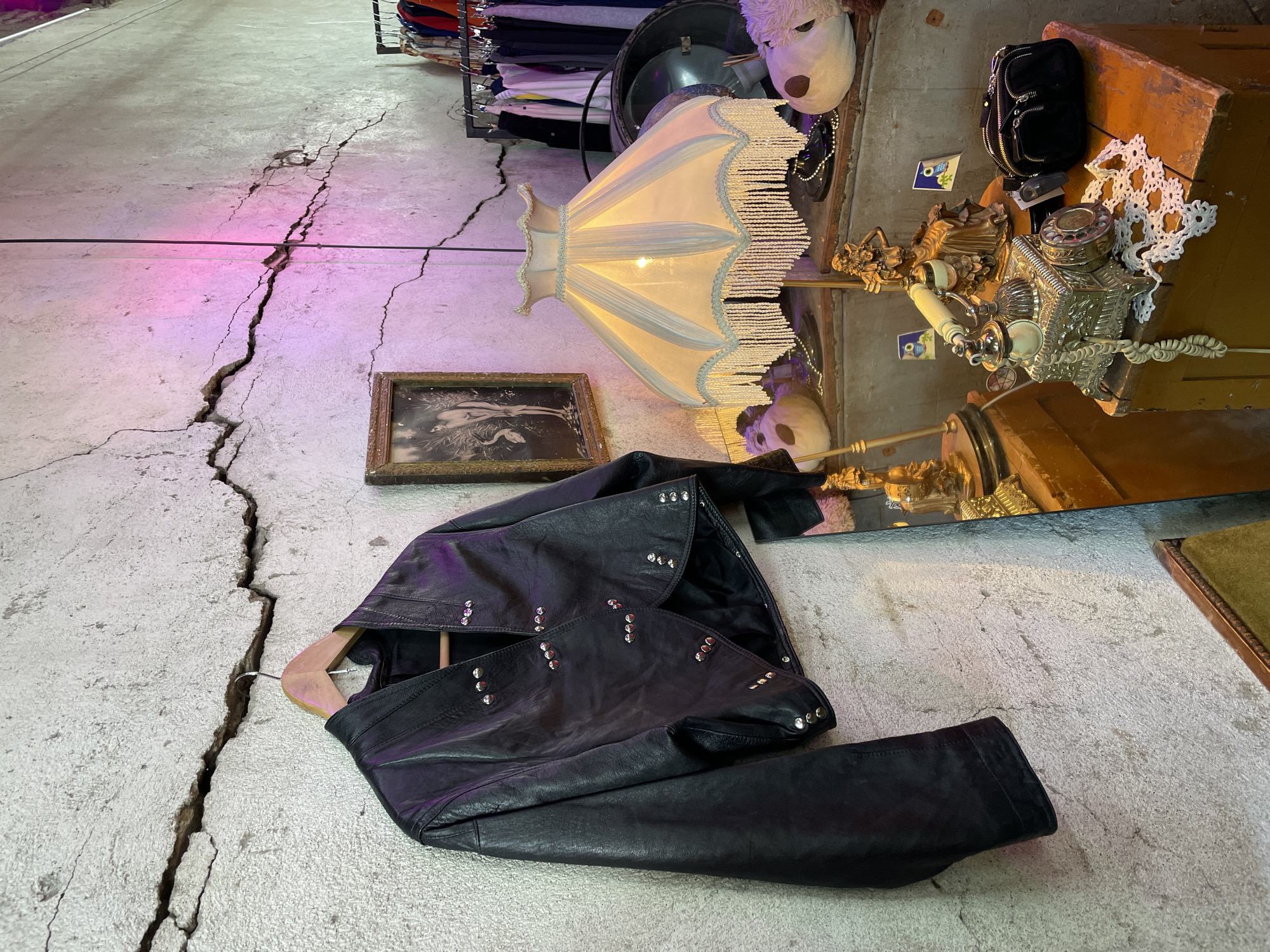 The image captures an indoor scene, possibly within a retail space, in Tbilisi, Georgia. The setting exhibits a cracked concrete floor, predominantly gray with pink hues, and several fissures running across its surface. A black leather jacket, adorned with studs, rests on a wooden hanger and lies partially on the floor. Adjacent to the jacket is a framed monochrome picture.
The scene also contains an ornate lamp with a fringed lampshade, a mirror reflecting the objects, and a display unit with various items including a silver tea set, decorative figurines, a black handbag, and a stuffed animal mask. In the background, a rack of clothes is visible.