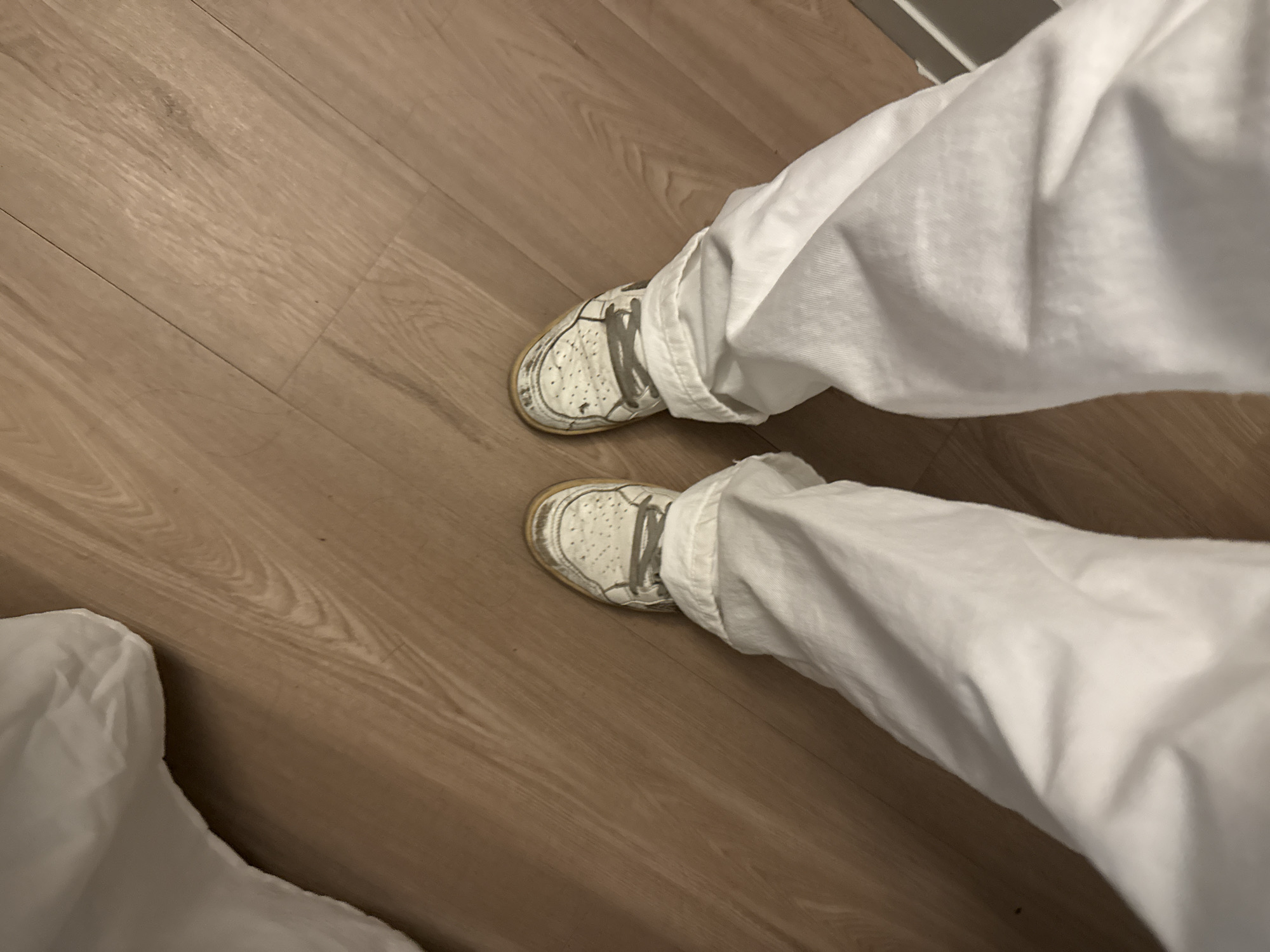 An overhead view captures a person's lower legs and feet, clad in white, loose-fitting trousers and white sneakers, standing on a light brown wooden floor. The trousers are casually wrinkled and appear to be made of a soft fabric. The sneakers feature grey laces and show noticeable signs of wear, particularly on their yellowish-brown soles and with some dark scuff marks on the white uppers.
The indoor setting is dominated by the wooden floor, which exhibits distinct grain patterns and visible seams between the planks. The floor has a subtle sheen, reflecting the ambient light. The lighting is soft and even, suggesting either diffused natural light or typical indoor illumination. No specific time of day or weather conditions can be determined. There are no other individuals or prominent objects in the frame, keeping the focus on the person's footwear and the floor. No visible text or branding is discernible on the clothing or shoes. While the context mentions Austin, United States, no specific visual cues within the image confirm this location.