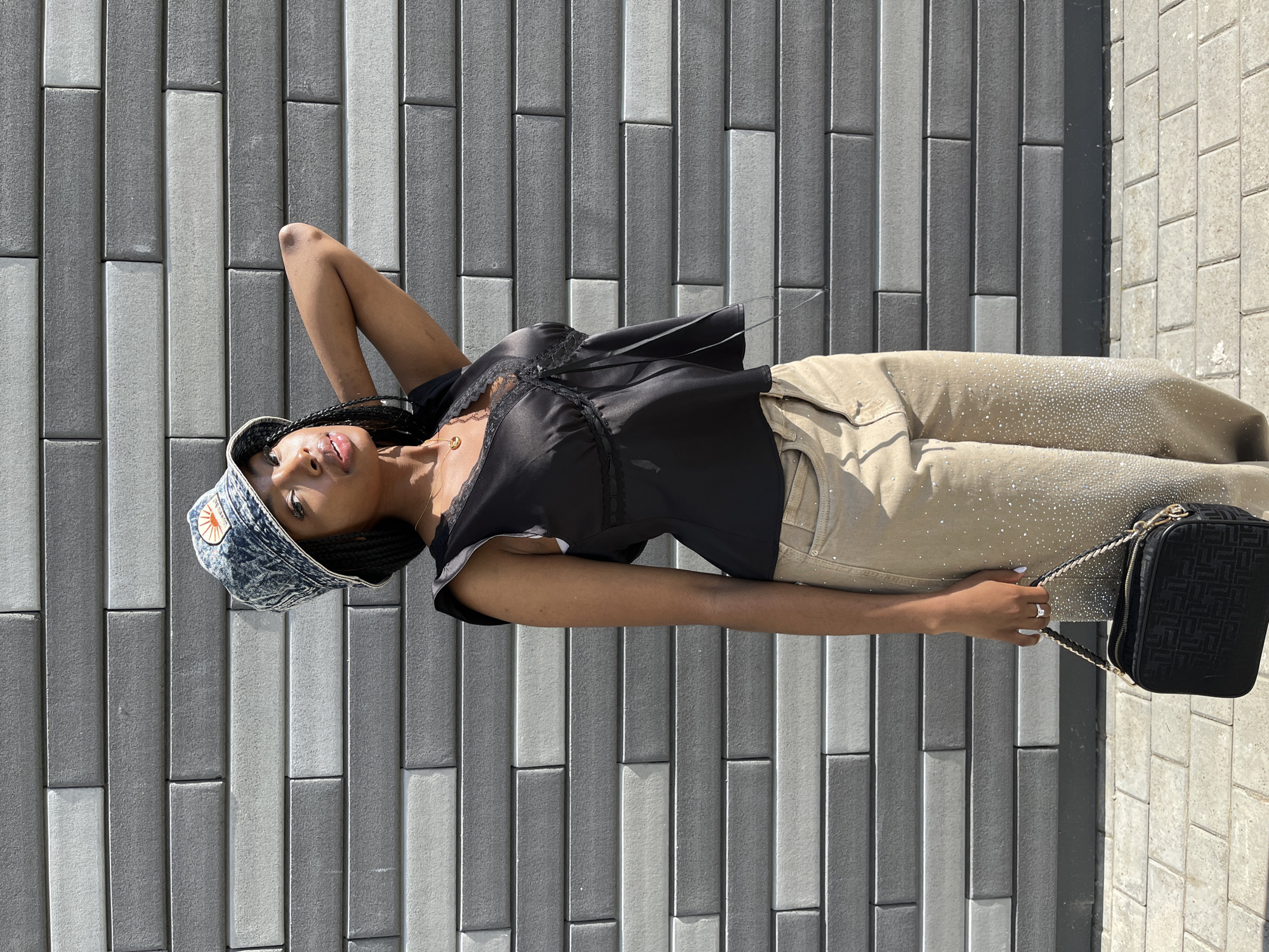 A young Black woman stands in front of a gray brick wall, her right arm raised and bent behind her head. She is wearing a denim bucket hat with a circular patch on the front, a black lace-trimmed top, and tan pants embellished with rhinestones. She holds a black handbag with a woven strap. The ground beneath her is paved with rectangular stones. The lighting suggests it is daytime. There is no indication that the location is Kazan, Russia.
