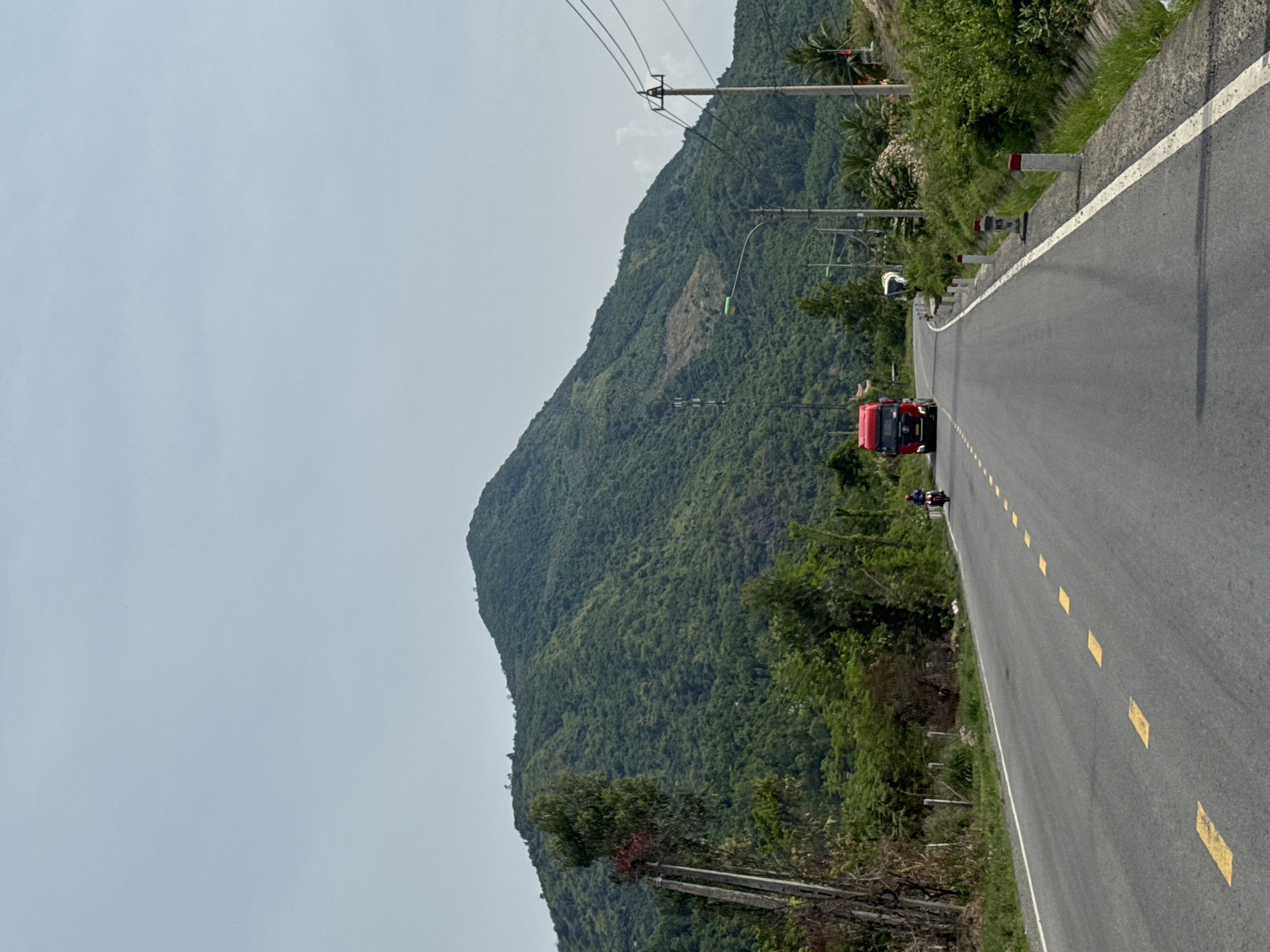 The image shows a scenic view of a winding mountain road in Nam Nha Trang Ward, Vietnam. A red truck is driving on the asphalt road, which is marked with yellow dashed lines. A person on a motorcycle is also visible on the road. The road is flanked by lush green vegetation on one side and a steep, tree-covered mountain on the other. Overhead power lines run along the side of the road. The sky is overcast, suggesting it might be a cloudy day. The overall scene conveys a sense of a journey through a natural landscape.