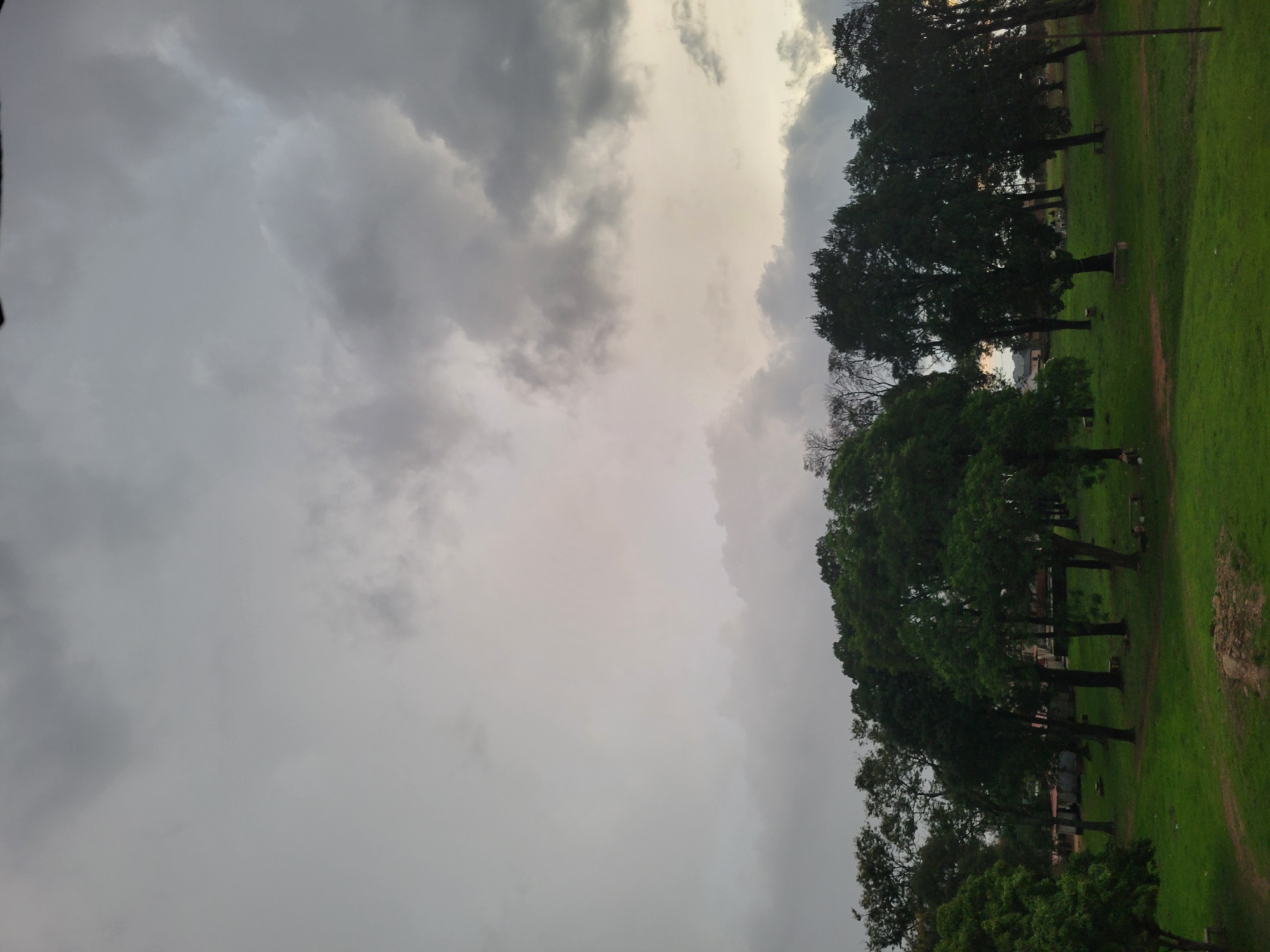 This image depicts a park-like scene within the city of Unknown, Unknown, under a heavily overcast sky. The upper two-thirds of the frame are filled with dense, stratified clouds ranging in color from light to dark grey, with some localized areas showing a faint lighter hue, possibly indicative of breaking cloud cover or twilight. The lower third of the image features a lush green grassy area, which appears slightly uneven with some visible dirt paths or patches. A cluster of mature trees with dark green foliage dominates the midground, forming a dense canopy. Several simple, low-lying benches or stone structures are visible at the base of various trees, scattered across the grassy expanse. In the background, partially obscured by the trees, are glimpses of indistinct low-rise buildings or structures. The overall lighting is soft and diffused, contributing to a subdued atmosphere. No people or overt activities are visible in the scene.