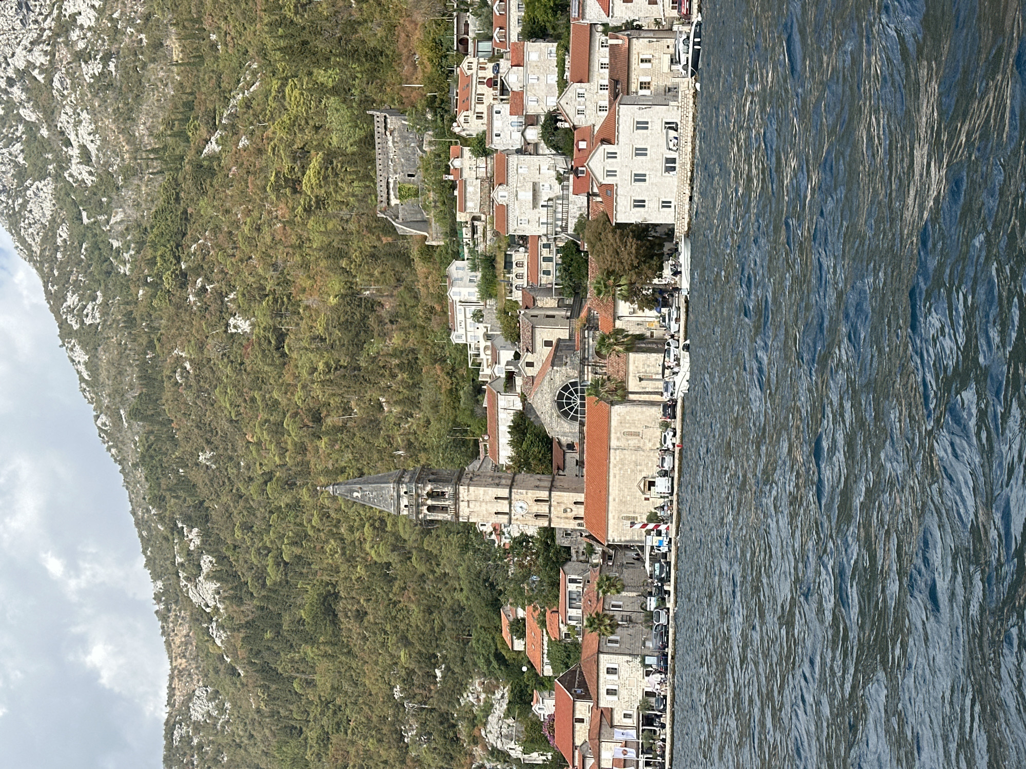 An eye-level, distant shot captures the waterfront of Perast, Montenegro, with the bay in the foreground and a steep, forested mountain range dominating the background. The water is a deep blue with visible ripples and waves. Along the shore, a dense cluster of stone buildings with red-tiled roofs forms the town. A prominent, tall bell tower with a clock face rises above the other structures, featuring arched openings and a pointed spire. To the right of the bell tower, a large building with a semicircular arched window is visible. Further up the hillside, ancient stone fortifications are integrated into the rocky terrain, with remnants of walls and a structure visible. The mountain slopes are covered in lush green trees, with patches of exposed rock and lighter-colored terrain higher up. A few vehicles are parked along the waterfront promenade. Several boats, including small motorboats and a larger yacht, are docked or moored in the bay. The sky is overcast with some lighter clouds.