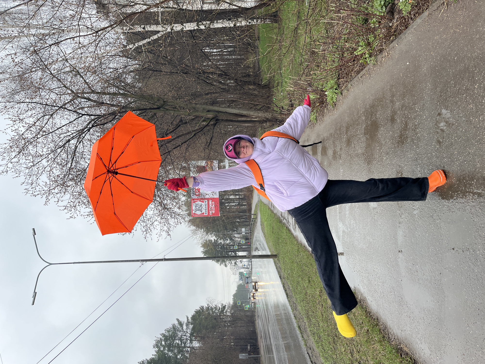A person, likely a young woman, poses playfully on a wet paved path in Chaikovsky, Russia, on an overcast day. She stands with arms outstretched and one leg lifted, giving the impression of flying or balancing. She wears a light purple puffer jacket, black pants, a pink baseball cap, and red gloves. Her attire is vibrant, featuring a bright orange open umbrella in her left hand, an orange rain boot on her right foot, and a contrasting yellow rain boot on her left foot. Orange backpack straps are visible over her shoulders.
The scene is an outdoor street or park area with a paved path and a road. The ground is wet, indicating recent rain or drizzle, and the sky is a uniform grey. Bare trees line the path and the road, suggesting late autumn, winter, or early spring. A grassy verge runs alongside the path. In the background, cars with headlights on drive along the road, and traffic signs are visible. A prominent lamp post with a "Y"-shaped top stands against the sky.
A red banner hangs from a tree in the mid-ground. Clearly visible text on the banner reads "12 лет РОСТИКС!", with smaller, less distinct text beneath it. The person's face conveys a joyful and spirited expression.